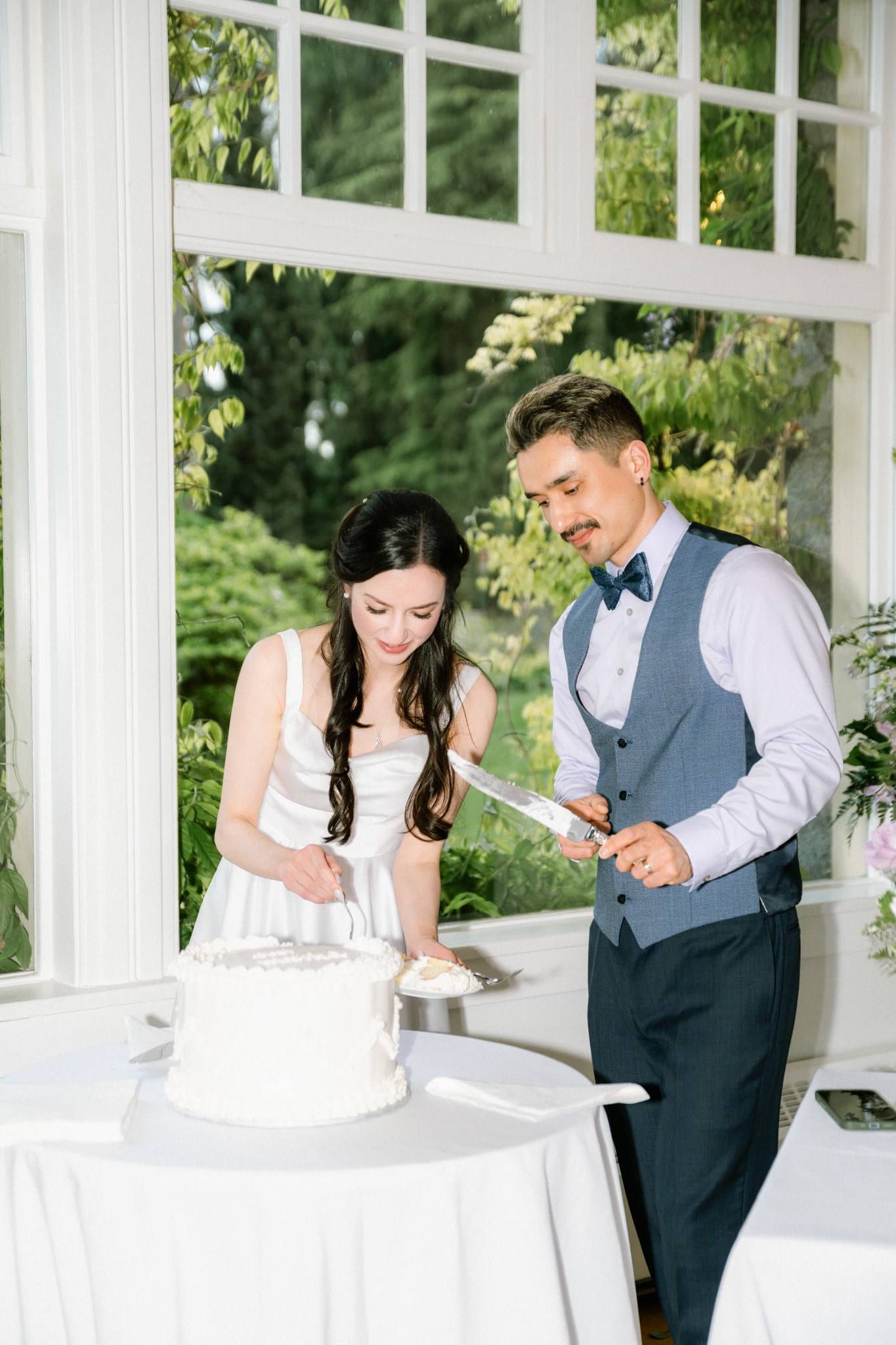 Bride and groom cutting their white wedding cake by a bright window