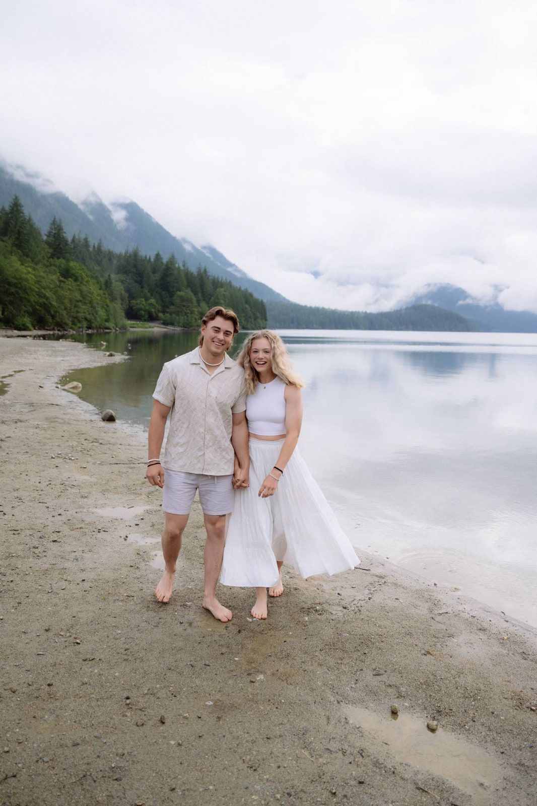 Lifestyle wedding photography, a couple stands barefoot and smiling on a sandy lakeshore with forested mountains behind