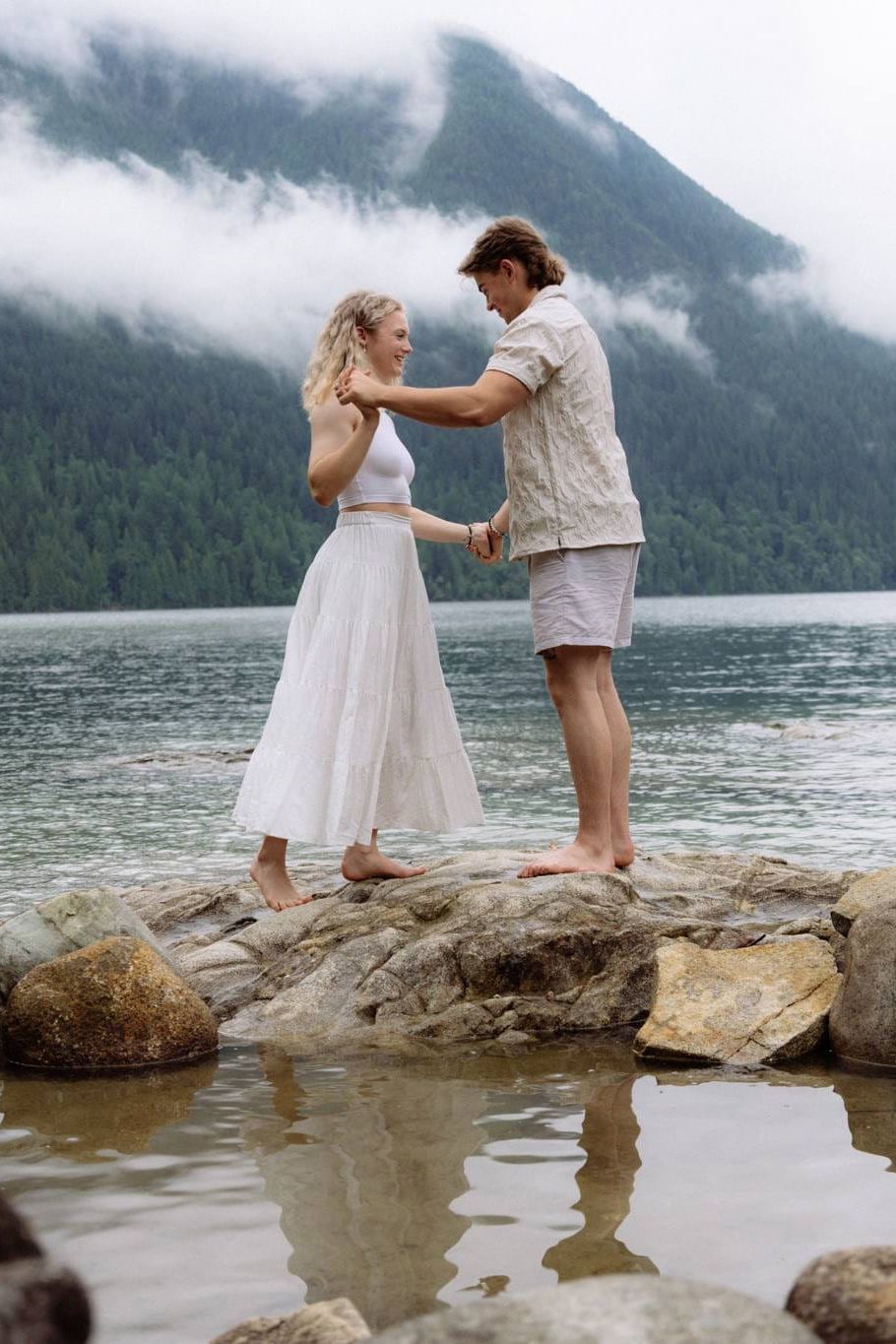 Professional couple photography over forested mountains, a couple holds hands on lakeside rocks with low clouds