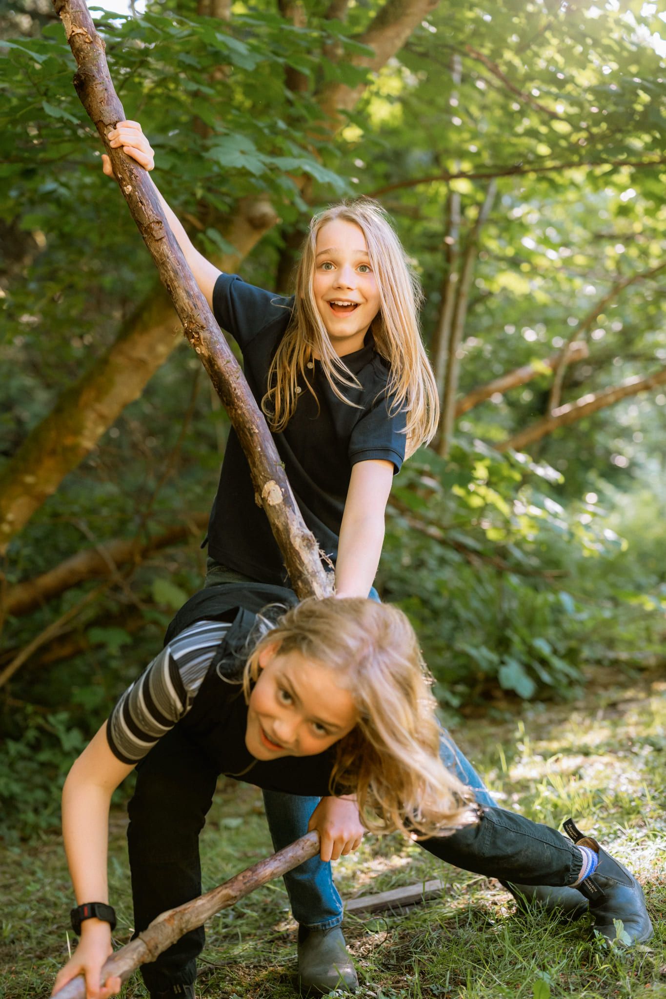 Forest family photoshoot, two sisters laughing as they play with a long stick