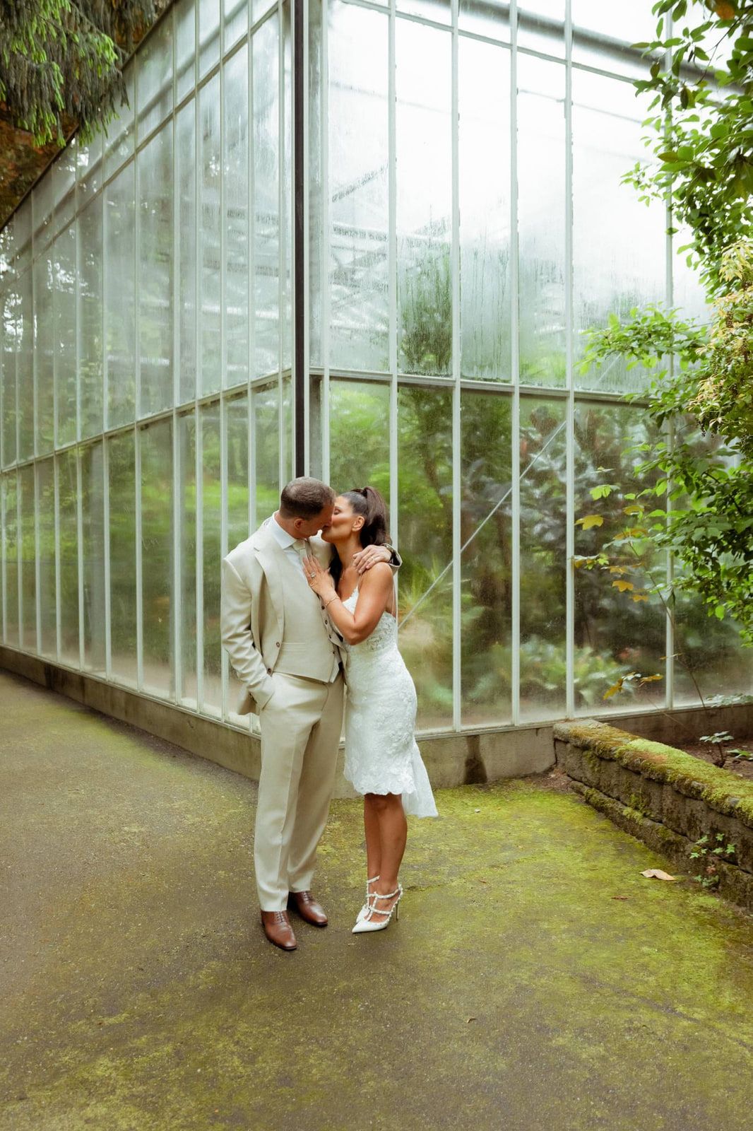 Garden engagement photos beside tall glass greenhouse walls, a couple in wedding attire kisses on a mossy path