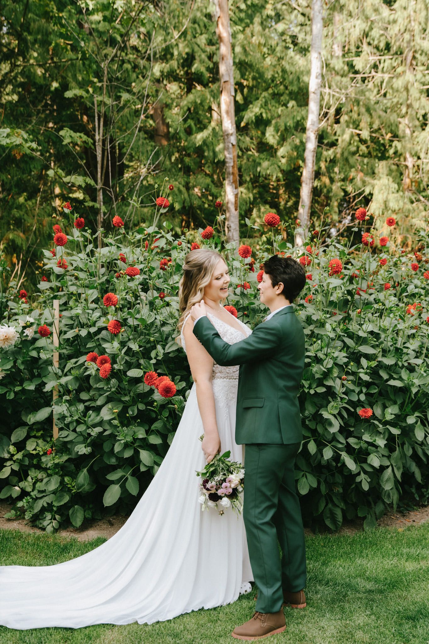 Intimate wedding photography, bride in a flowing white gown and groom in a green suit stand face to face among red dahlias in a garden, bouquet in hand
