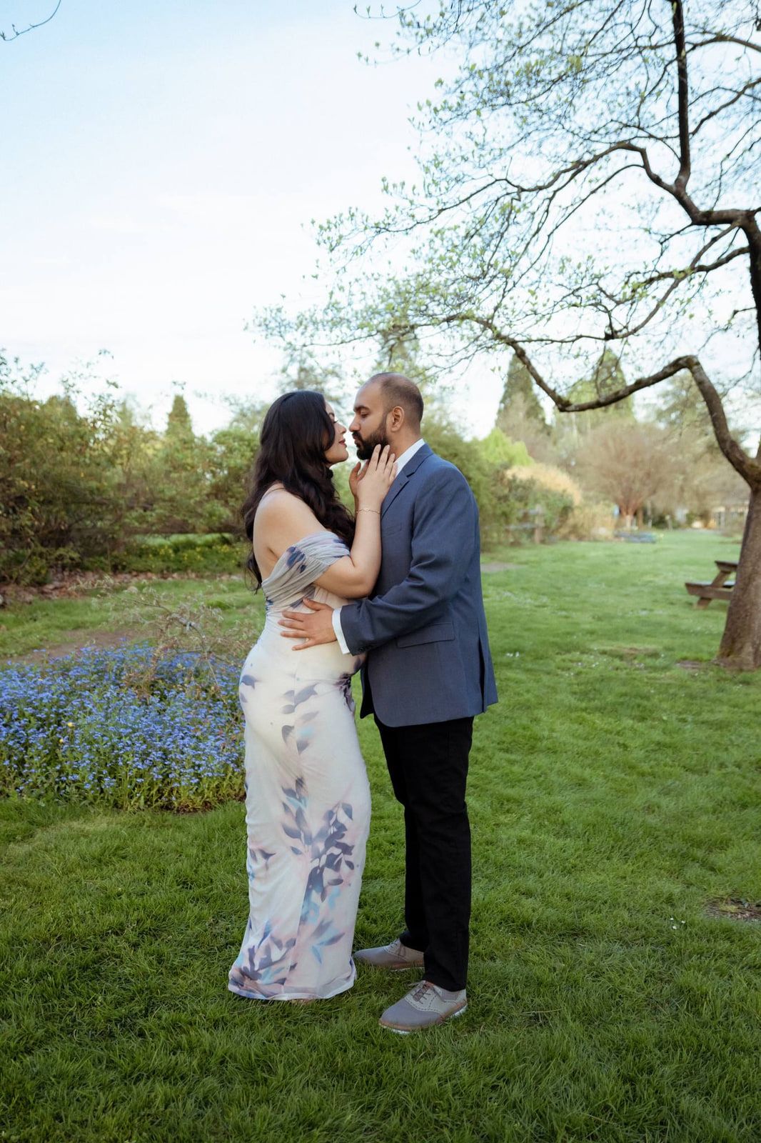 Lifestyle engagement photos, a couple shares a kiss on a lawn beside a bed of blue flowers, one in a floral maxi dress