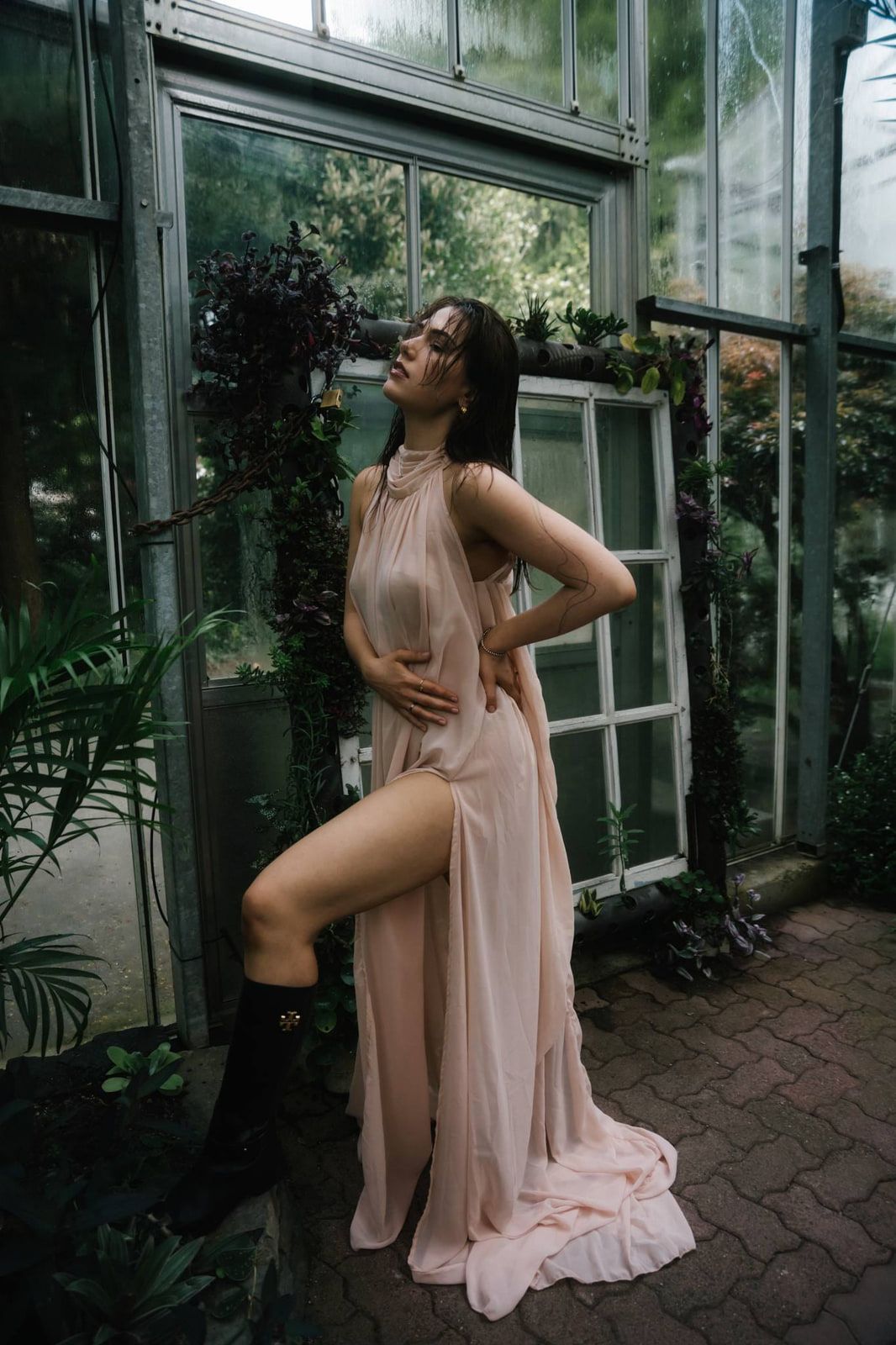 Natural portrait photography, a woman in a blush gown and black boots poses against an old window frame in a greenhouse