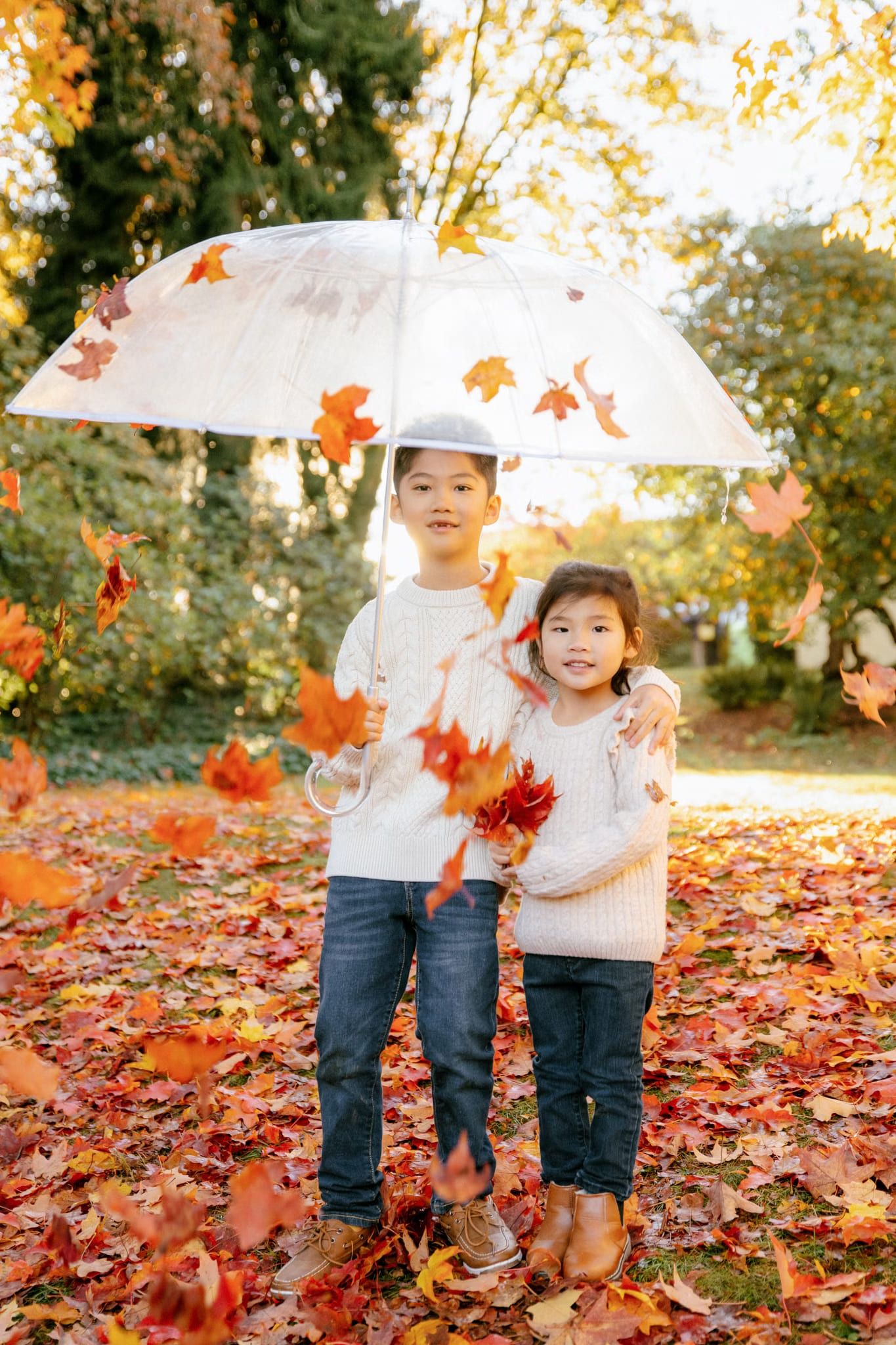 Two children in cream sweaters under a clear umbrella as autumn leaves fall