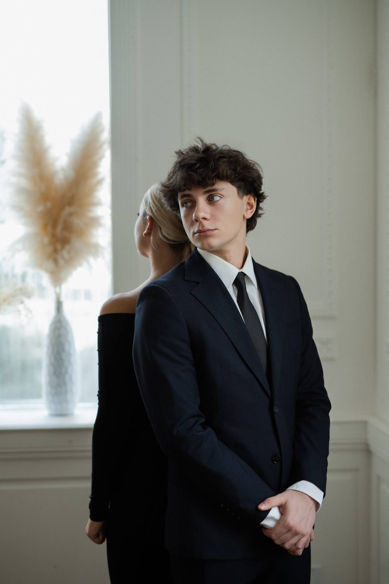 Indoor wedding photos, a couple by a tall bright window, he in a dark suit looking over his shoulder, she in black resting against him