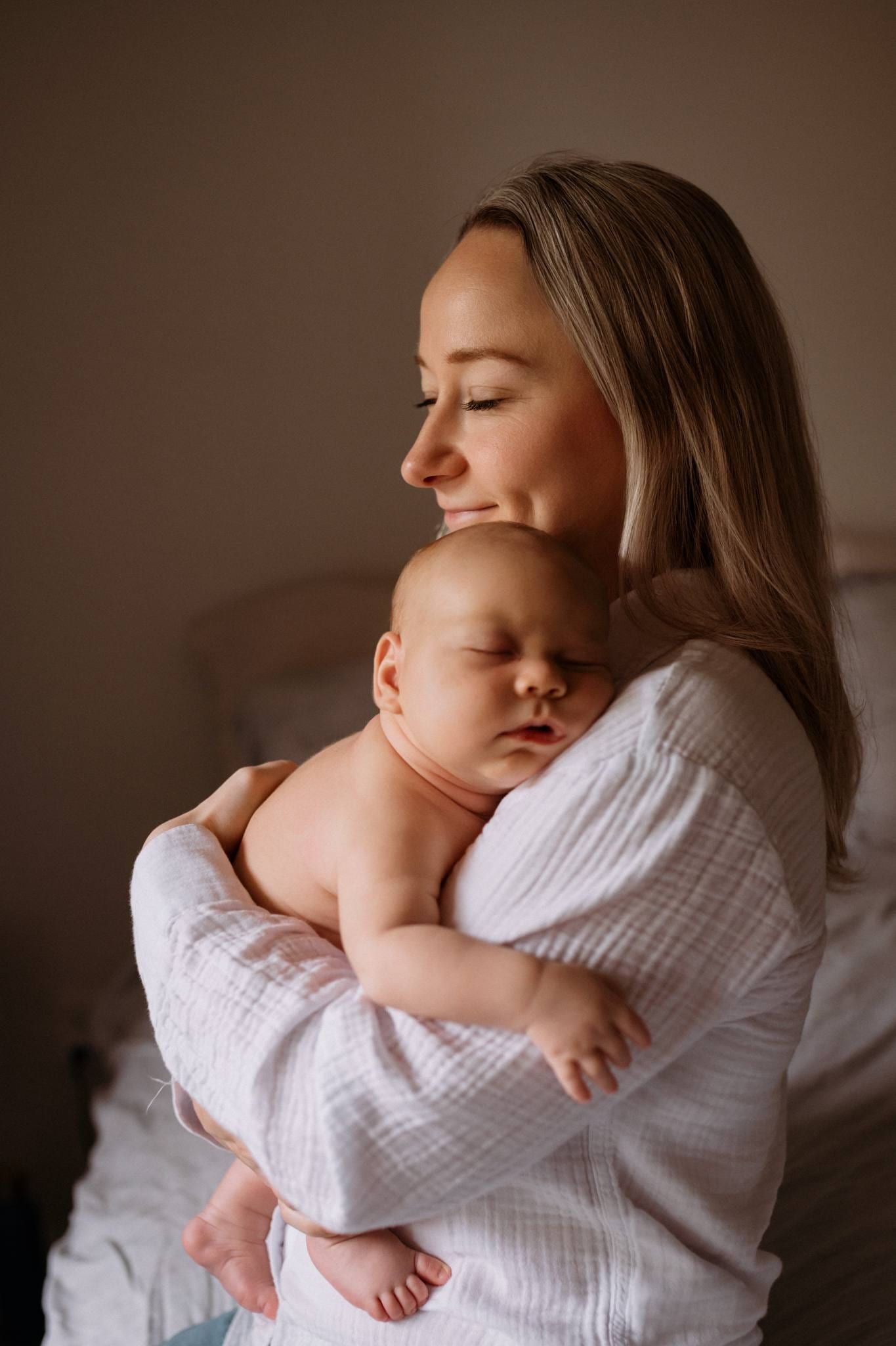 Mother holding her swaddled newborn against her shoulder in soft window light