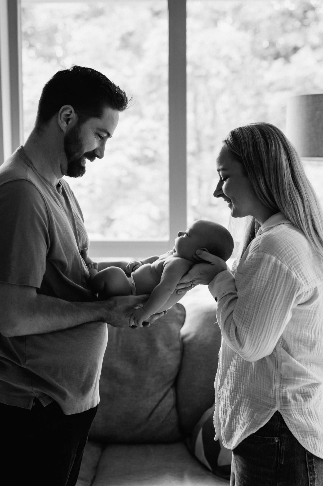 Black-and-white photo of parents holding their newborn between them by the window