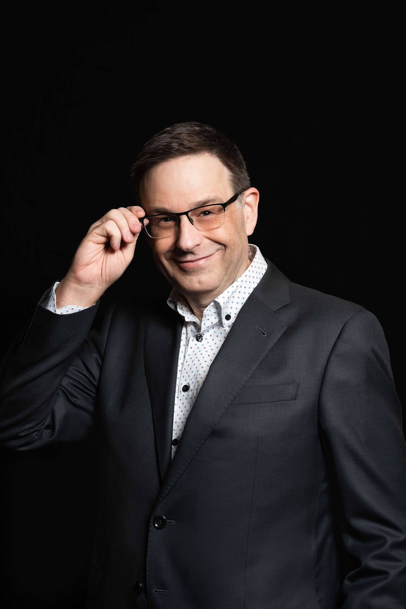 Professional headshot photoshoot, man in glasses and a dark suit adjusting his frames on a black backdrop