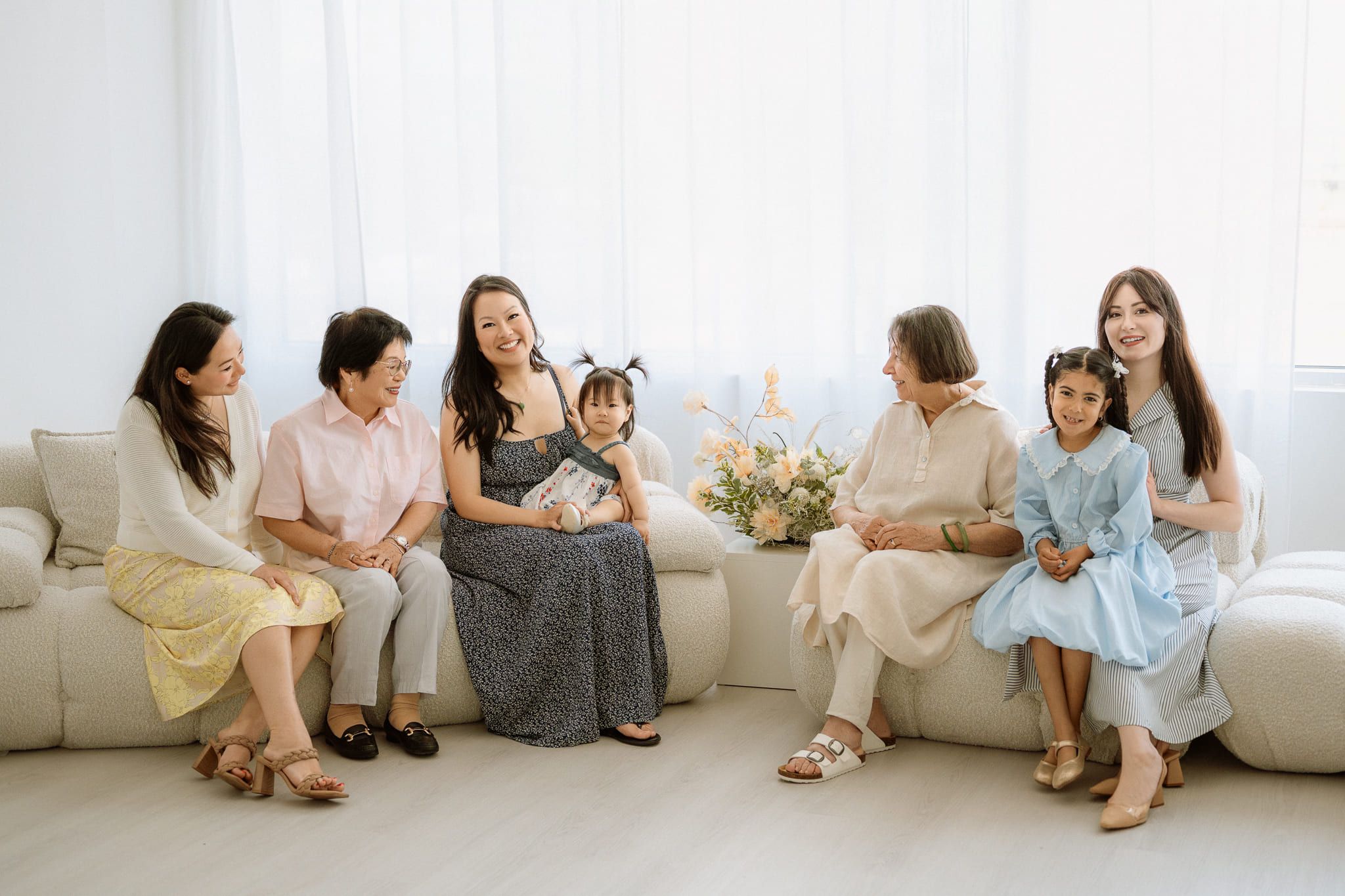 Family photo in home of three generations of women and girls smiling on a cream sofa by sheer curtains