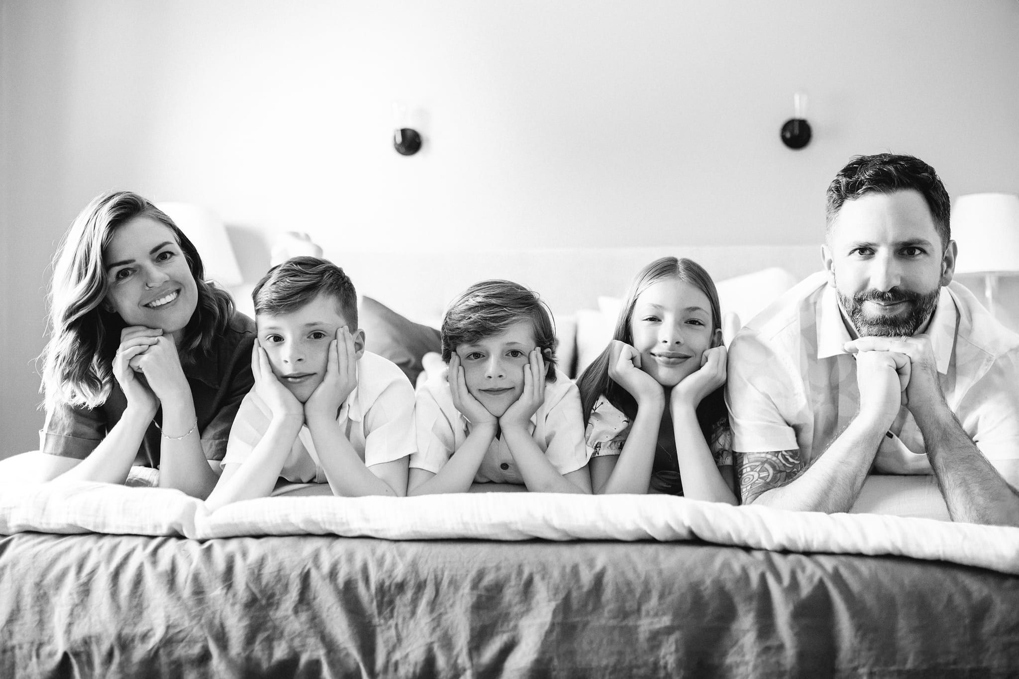 Home family photoshoot, mom, dad and three kids lying on a bed with their chins in their hands, in black and white