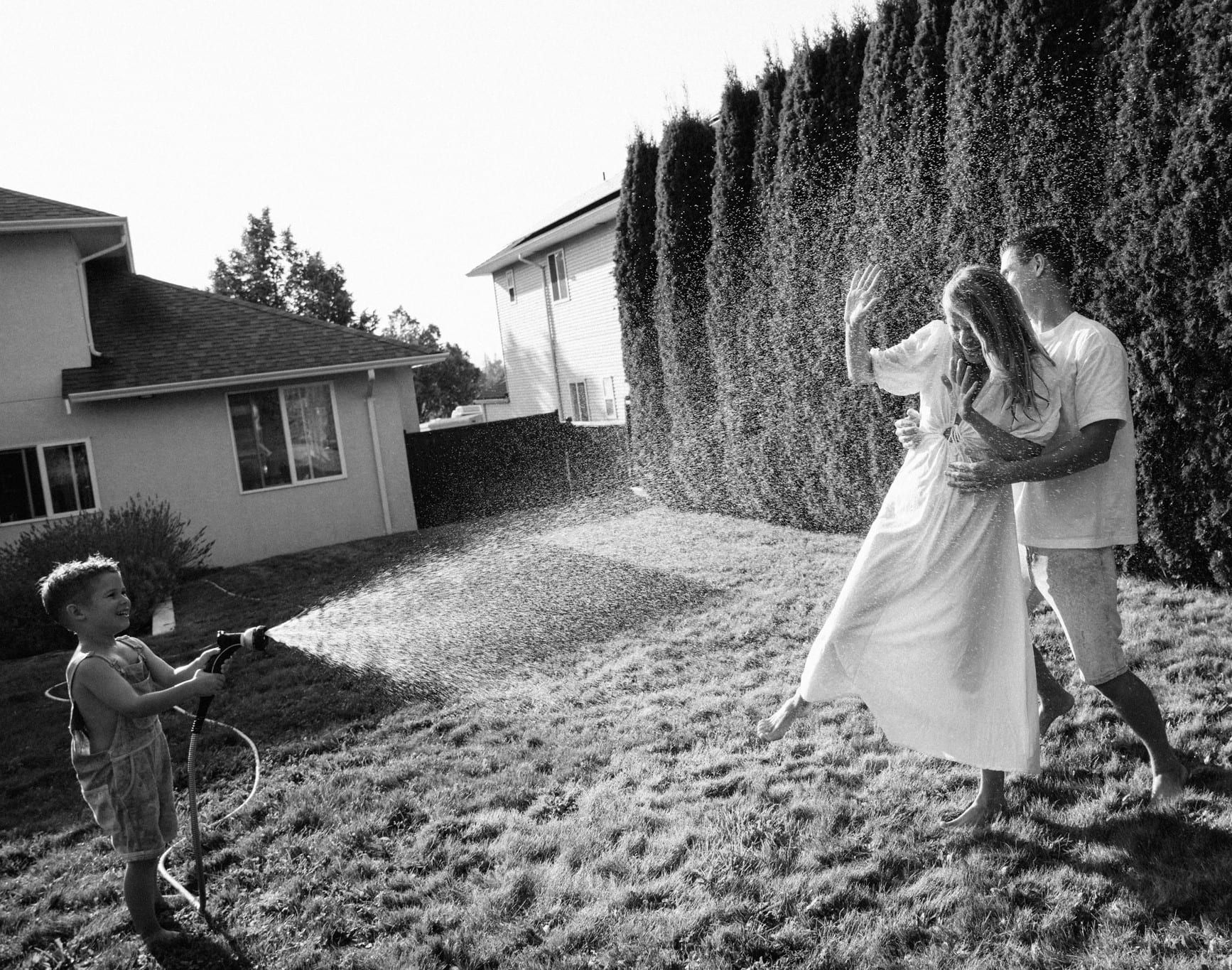 Black-and-white photo of a boy spraying his parents with a garden hose in a backyard