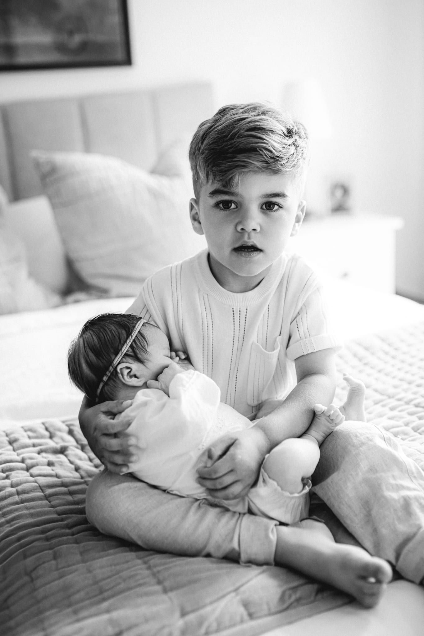 Newborn sibling pictures, big brother holding his newborn sibling on the bed, in black and white