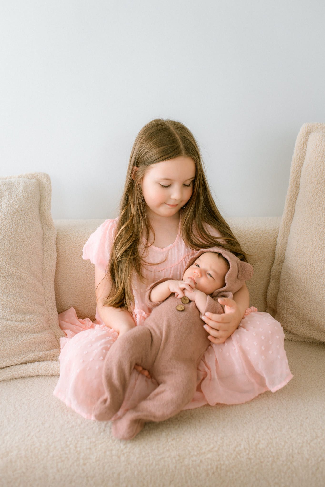 Newborn sibling photos, big sister in a pink dress holding her newborn sibling on a sofa