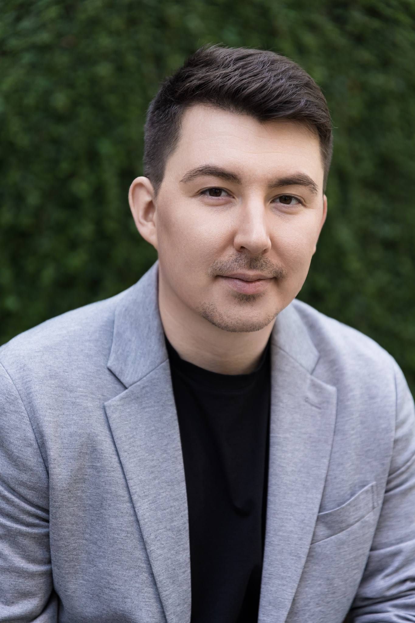Man in a grey blazer against a green hedge