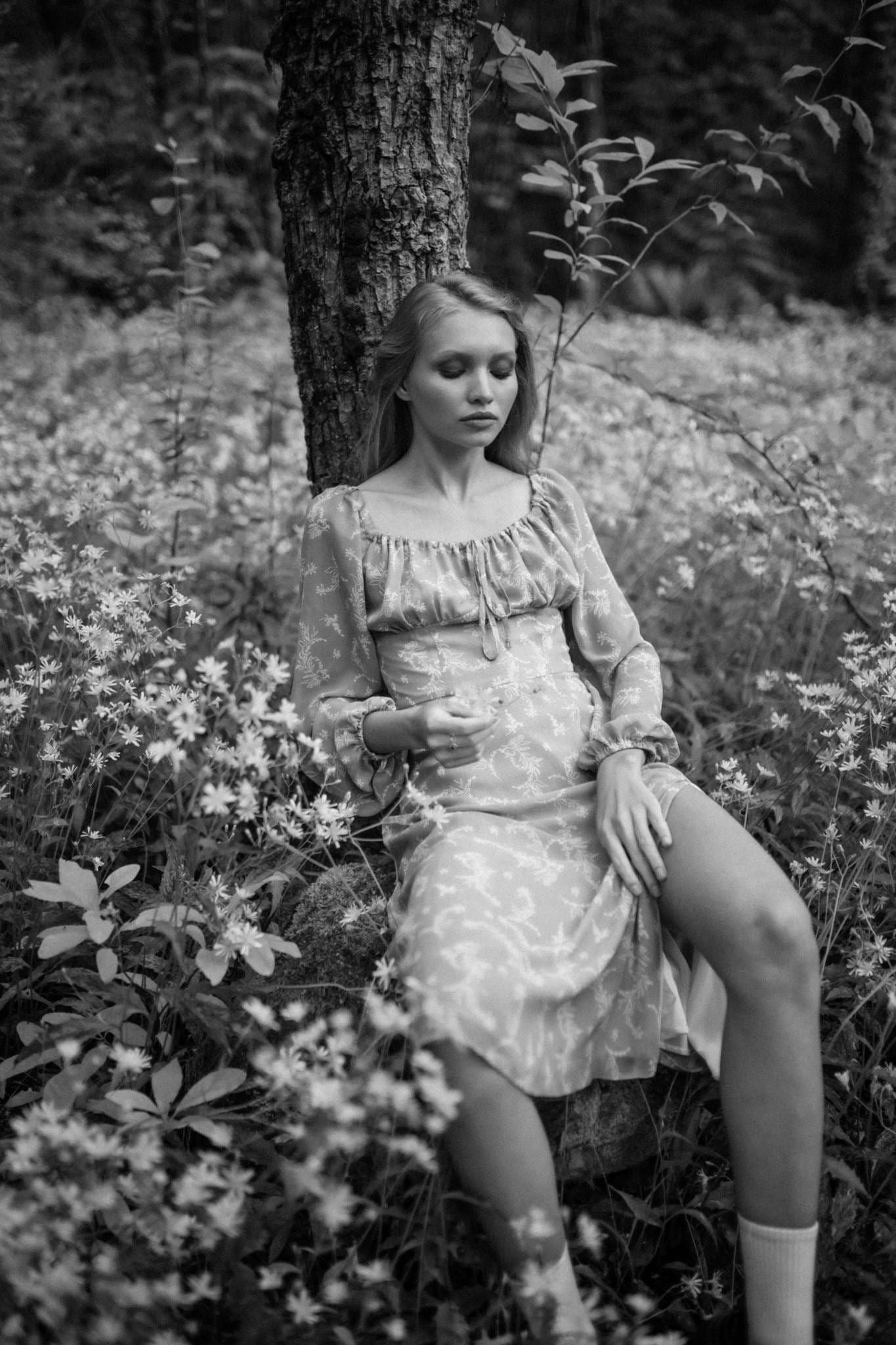 Black-and-white photo of a model in a floral dress seated at the foot of a tree