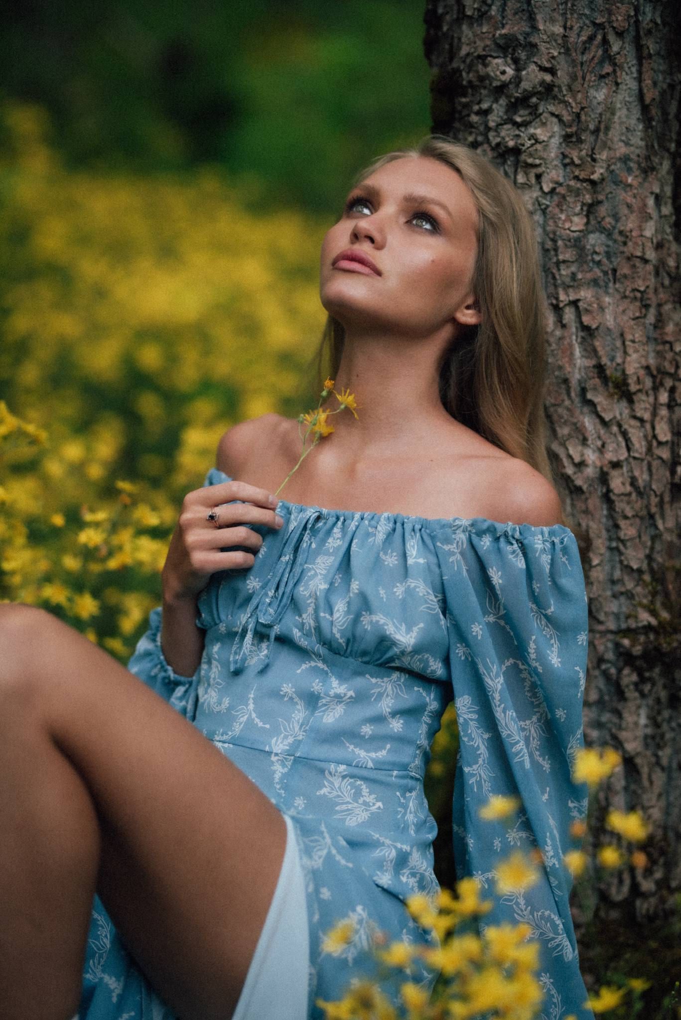 Model in a blue floral off-shoulder dress beside a tree among yellow wildflowers