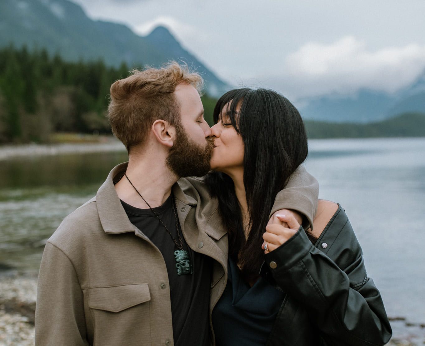 Engagement photos, couple kissing by a lake with mountains behind