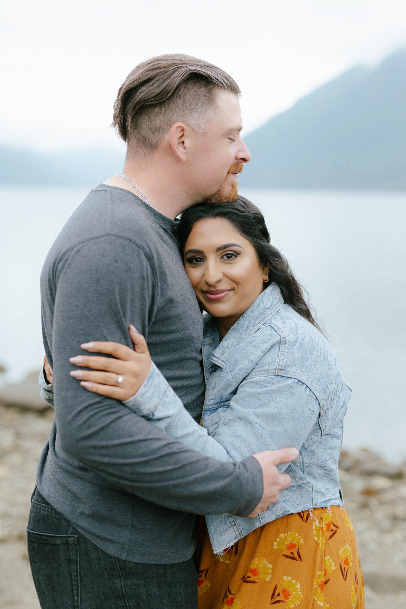Lake engagement photos, couple hugging cheek to cheek by a lake with mountains behind