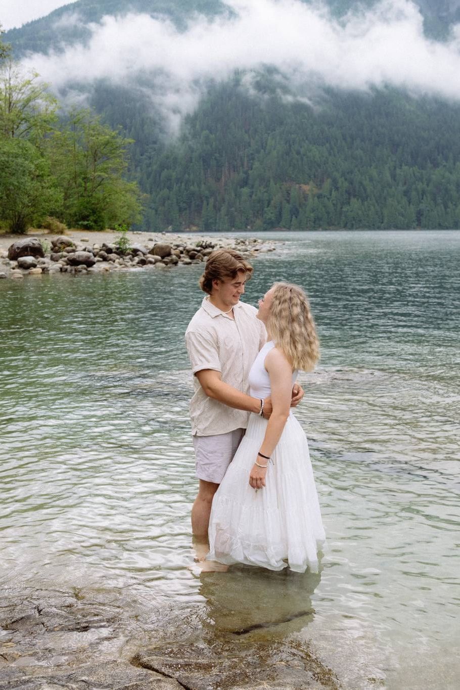 Classic engagement photos beneath cloud-covered mountains, a couple embraces while standing in shallow lake water