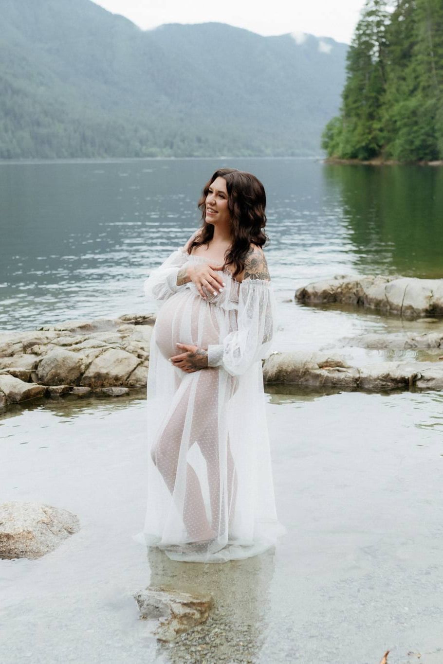 Outdoor pregnancy photos, an expectant mother in a sheer white gown stands in shallow lake water among smooth rocks