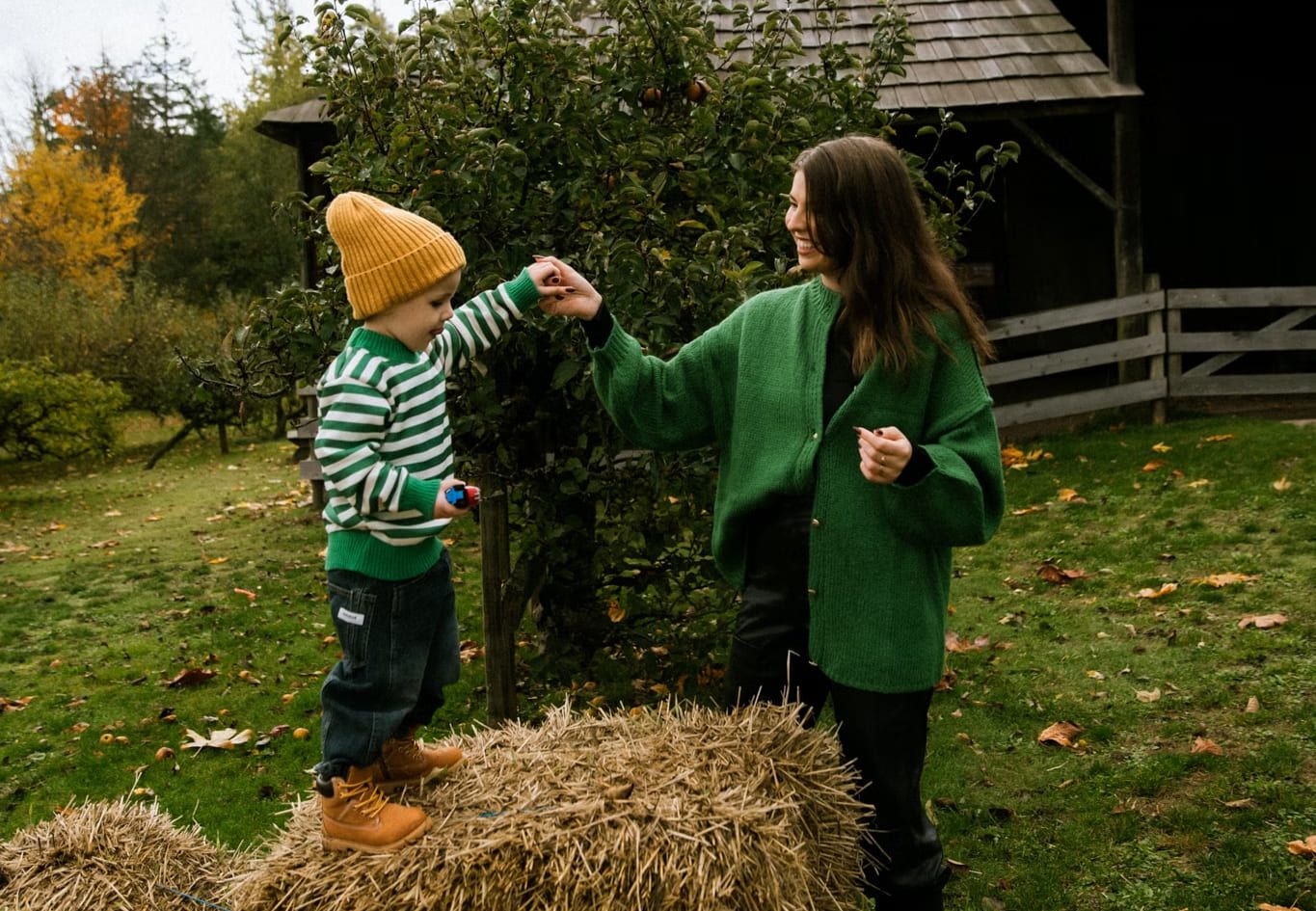 Mother in a green cardigan holding hands with her son as he stands on a hay bale in an autumn orchard