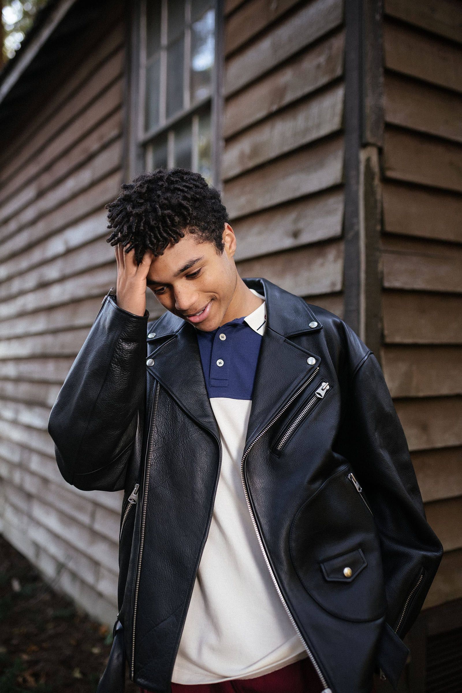 Young man in a leather jacket and white tee leaning against a wooden wall