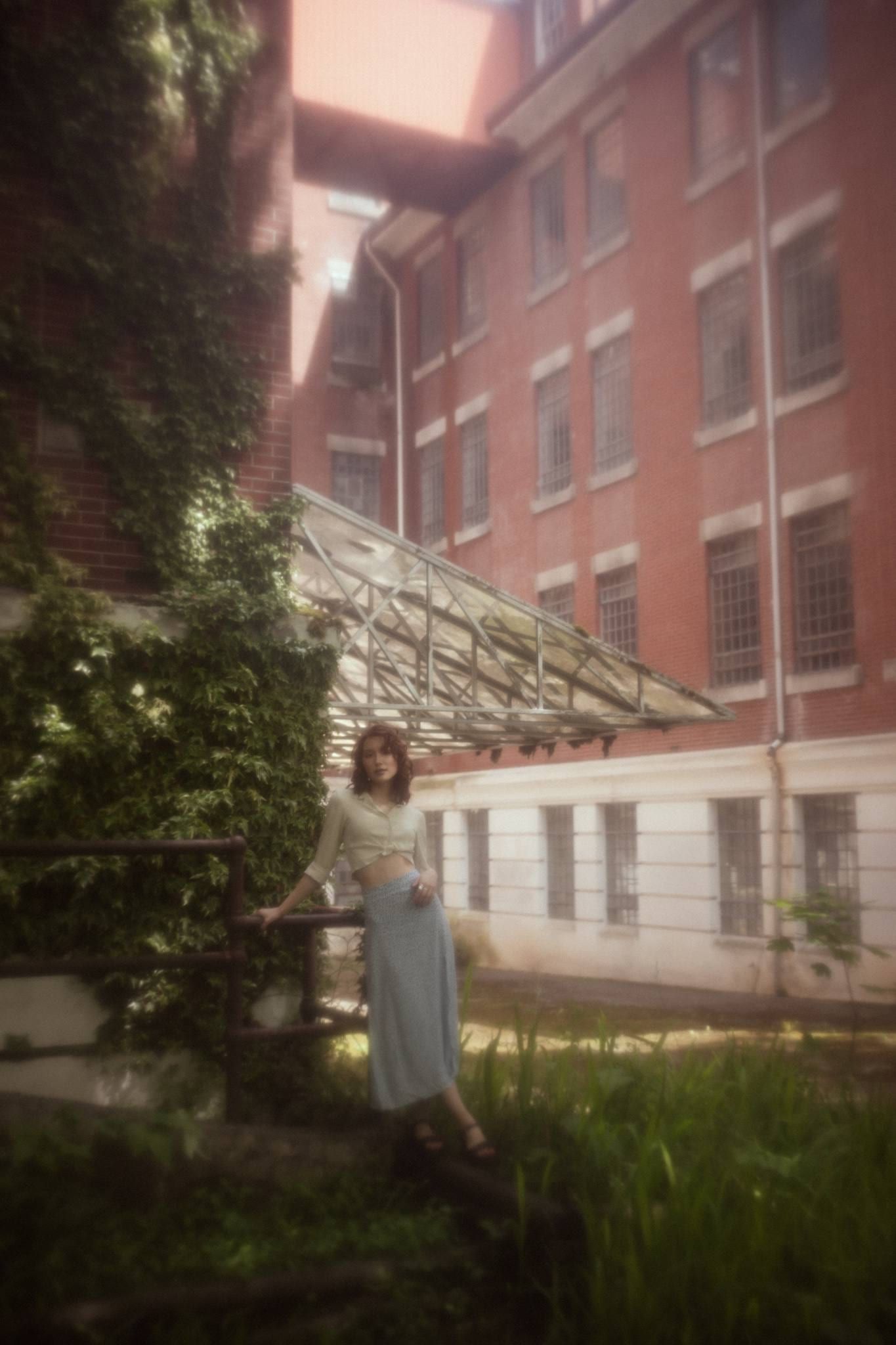 Model in a patterned dress standing in tall grass before a brick school