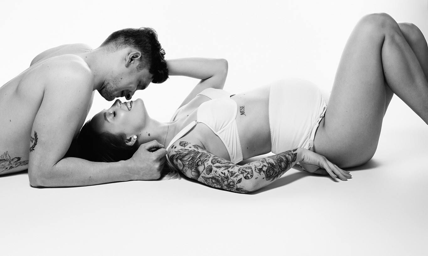 Black-and-white studio portrait of an expectant couple lying together, her tattooed arm across her bump