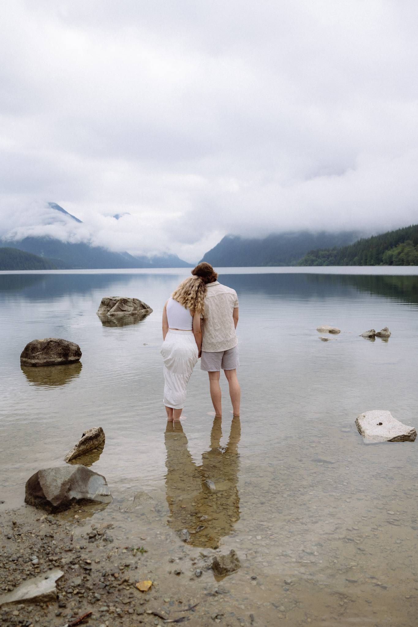 Couple standing ankle-deep in a still mountain lake beneath low cloud