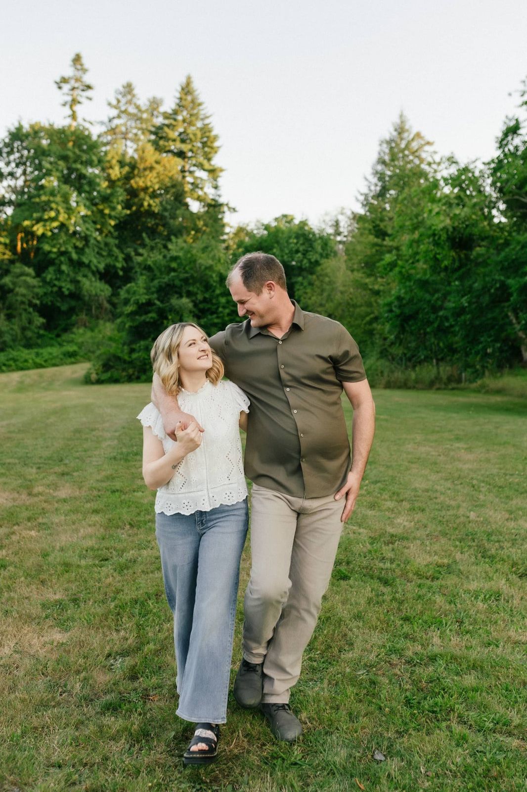 Engagement photos outdoors, a couple walks arm in arm across a grassy meadow edged with tall evergreens