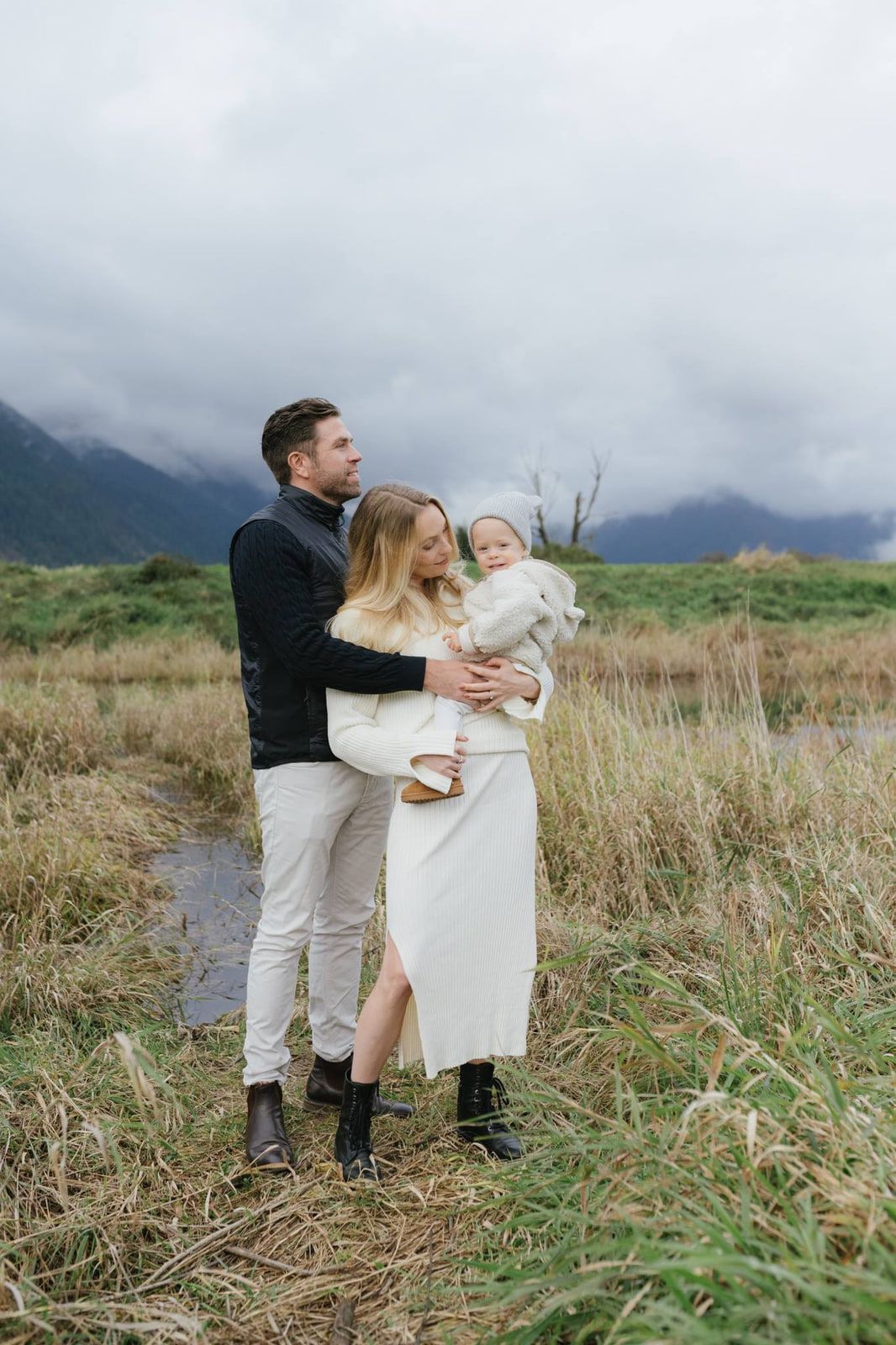 Professional family portrait photography, a couple stands in tall grass holding a toddler with a marsh and cloudy mountains behind