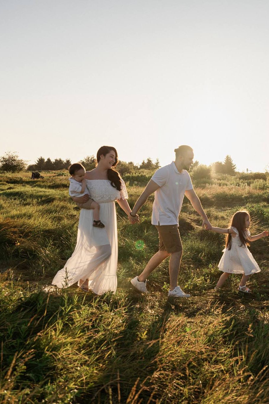 Family photo outside, a family of four walks hand in hand through tall grass in low evening sun
