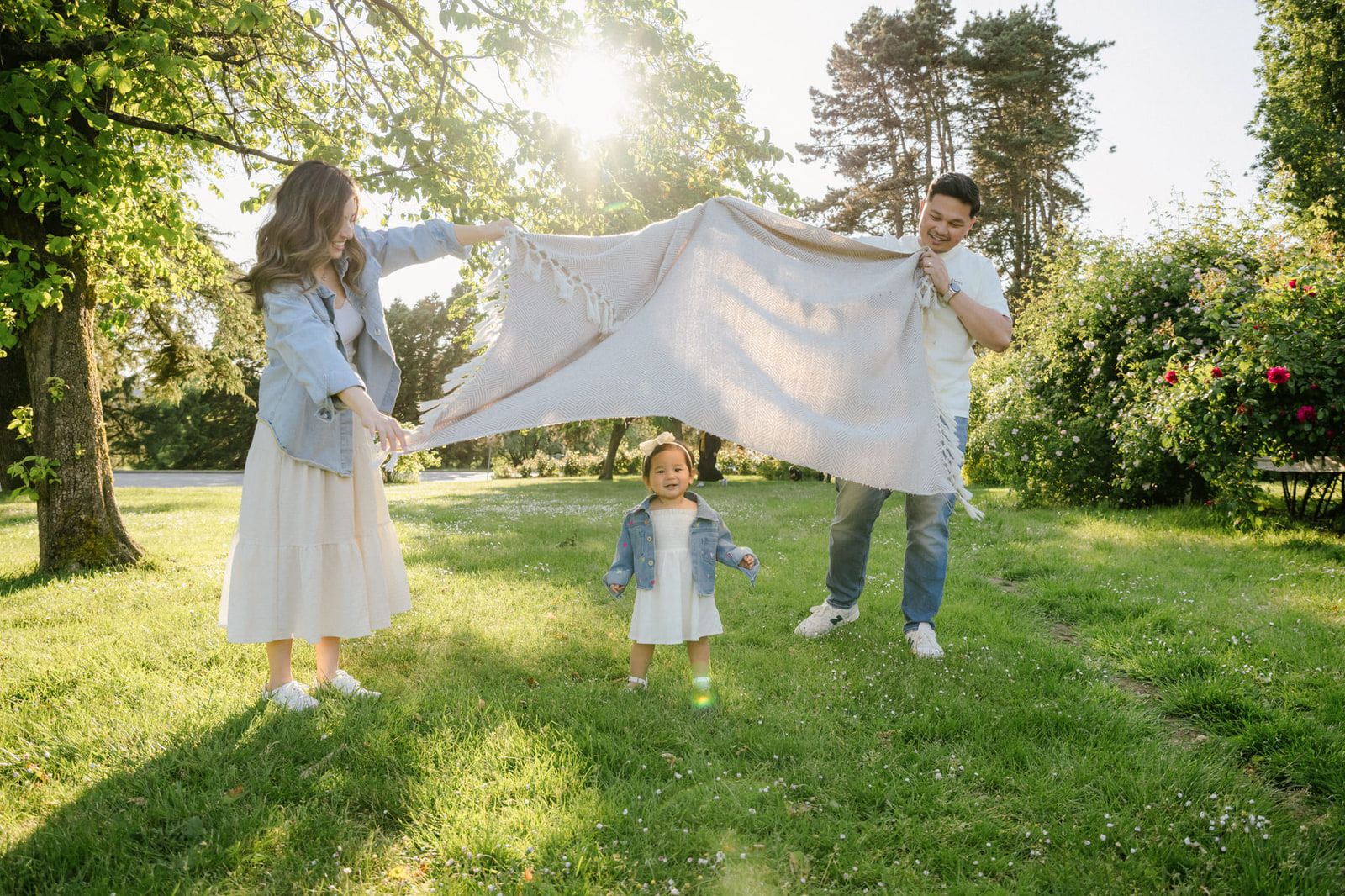 Coquitlam family photos, parents waving a white blanket in the sun as their little girl watches in a meadow