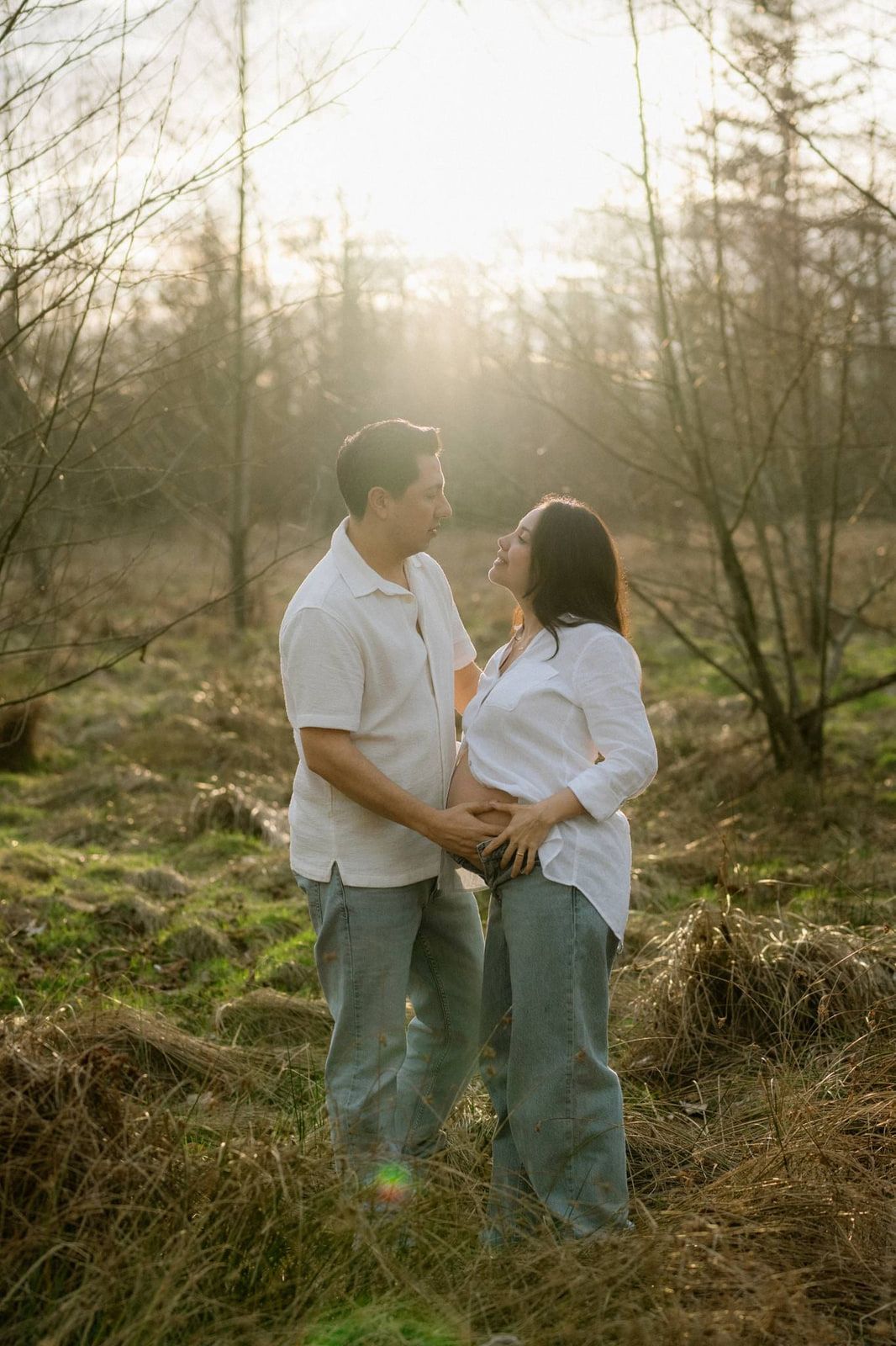 Maternity photos outside in backlit tall grass, a couple faces each other, hands resting on her bump