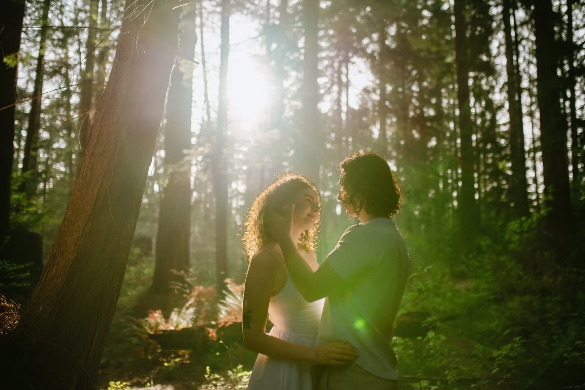 Couple gazing at each other in a sunlit forest with light flaring through the trees