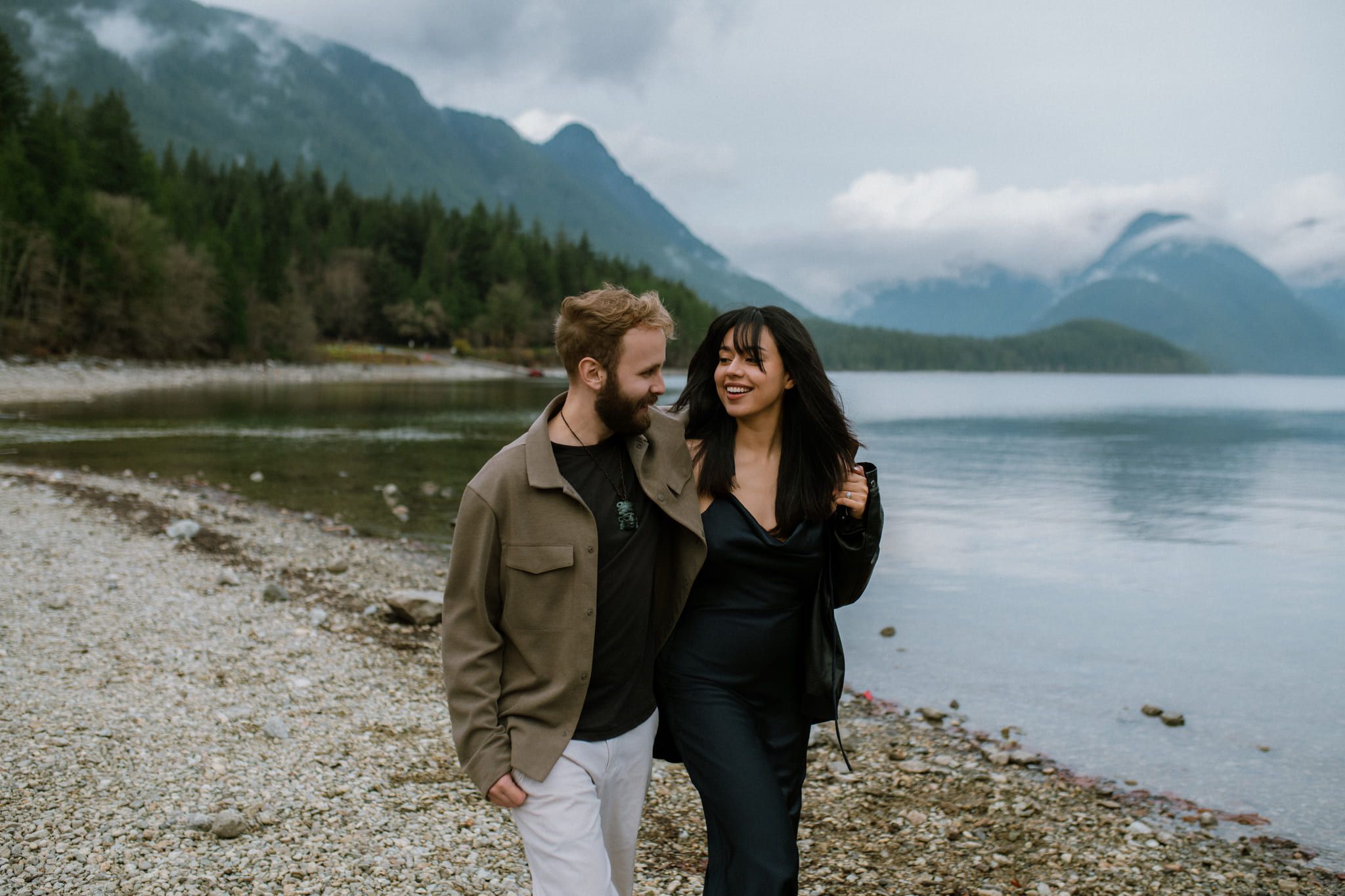 Engagement photos below misty mountains, couple laughing as they walk along a lakeshore