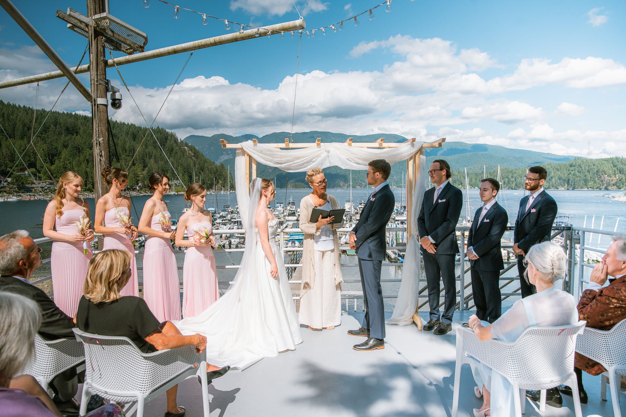 Natural wedding photography, ceremony on a waterfront deck with the bridal party in pink and the mountains behind