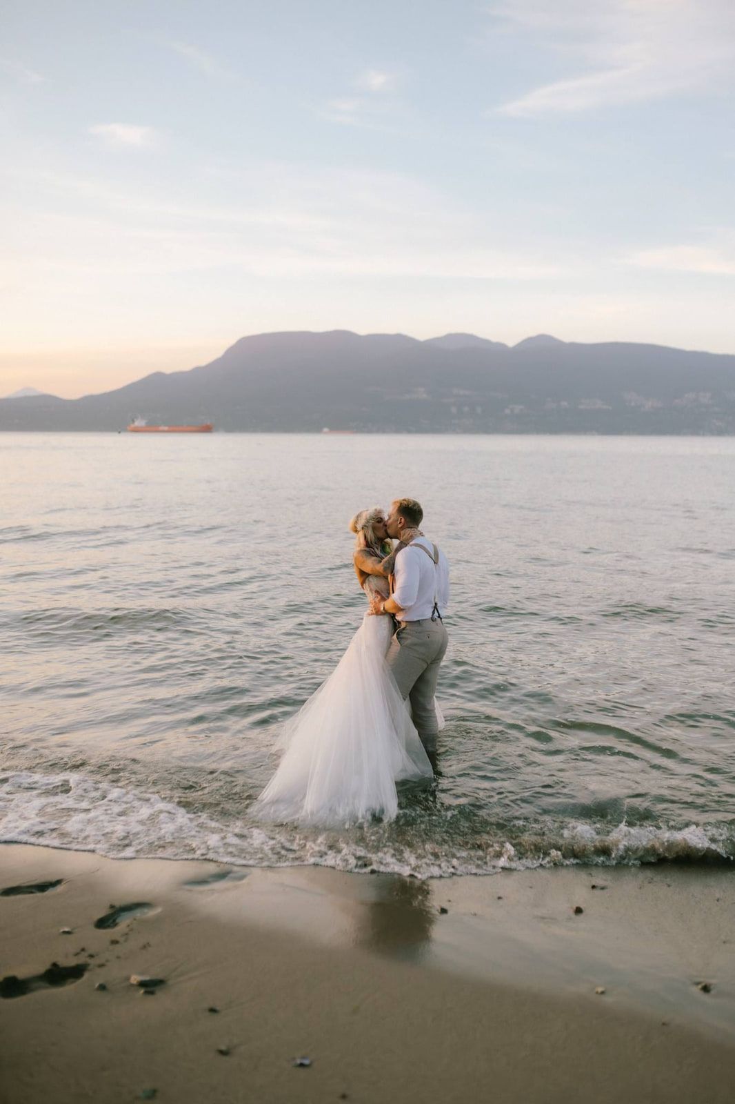 Mountain elopement photos across the water, a couple in wedding attire embraces in the shallow waves with mountains