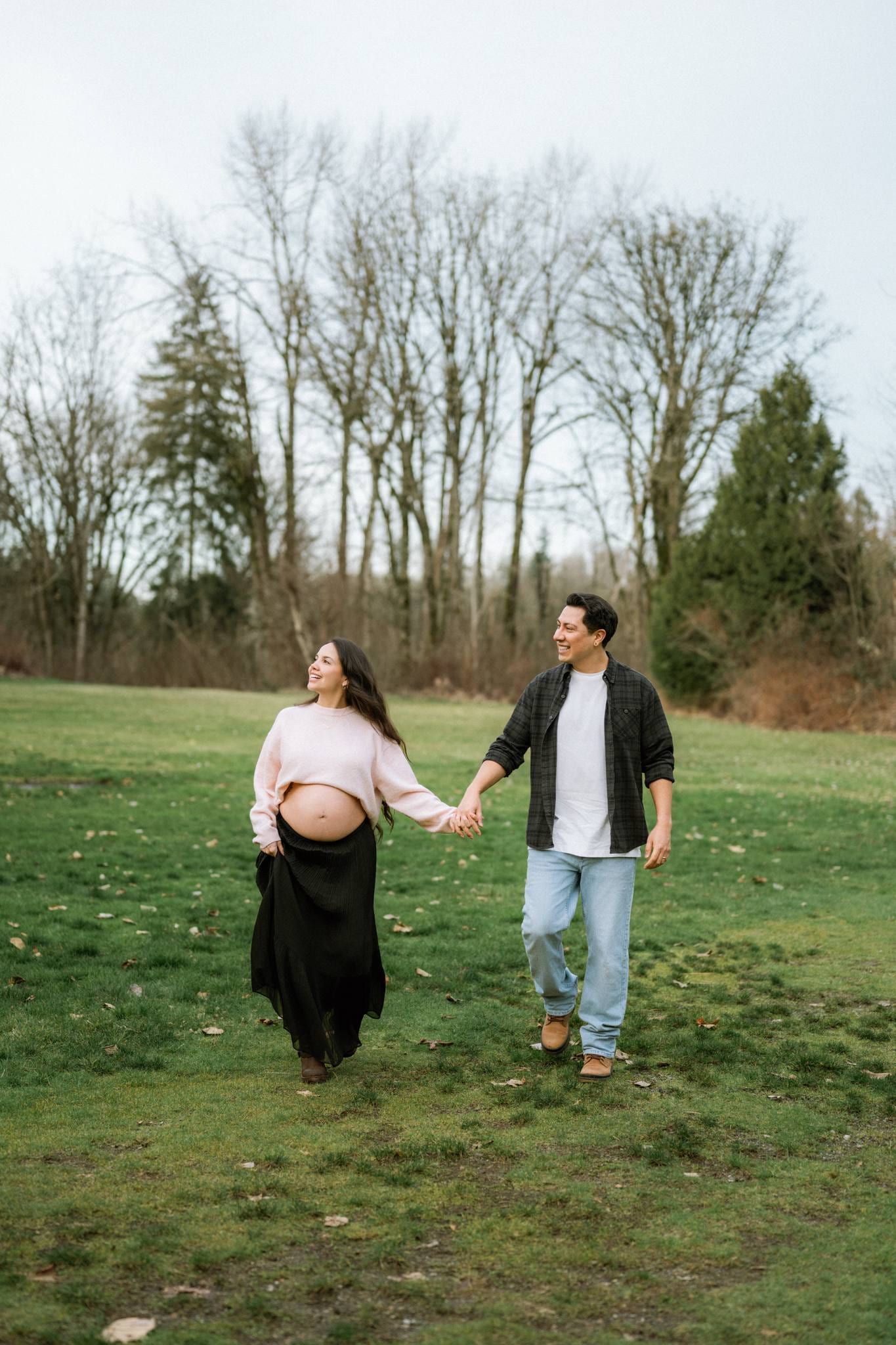 Expectant mother in a pink sweater walking hand in hand with her partner across a park lawn