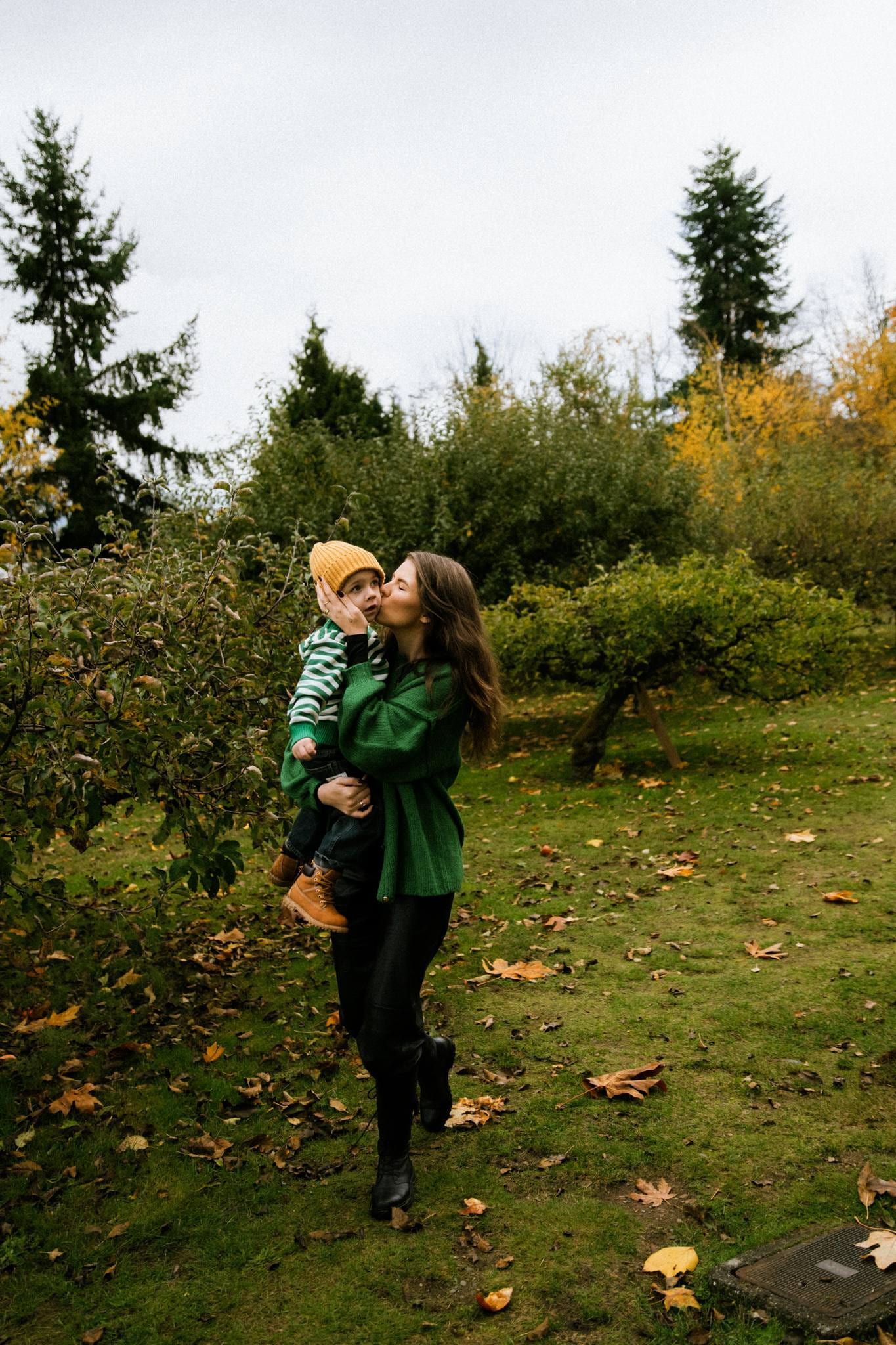 Mother in a green cardigan kissing her son in a yellow beanie among autumn apple trees