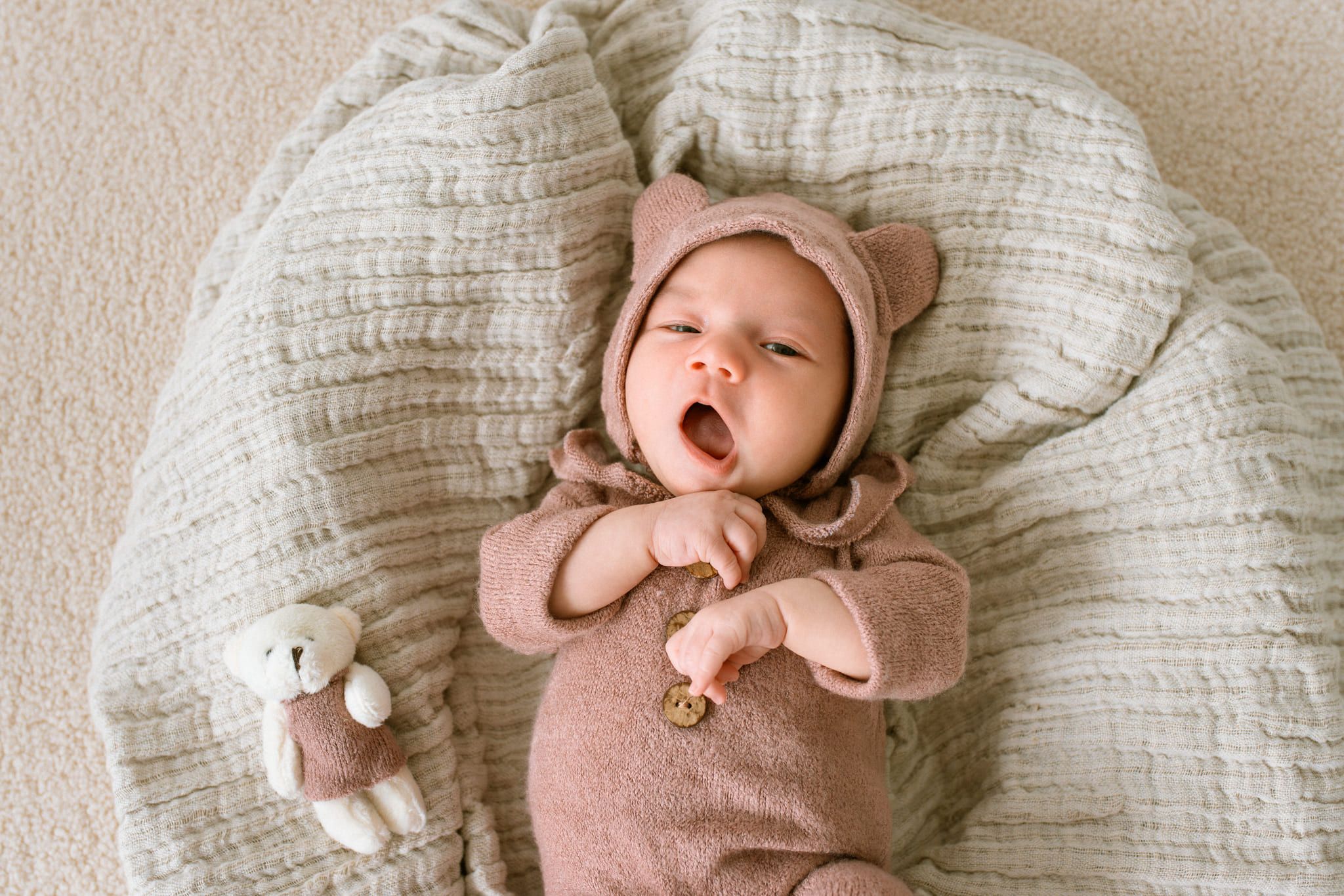 Newborn shoot, newborn in a pink knit romper and bear hood, yawning on a cushion