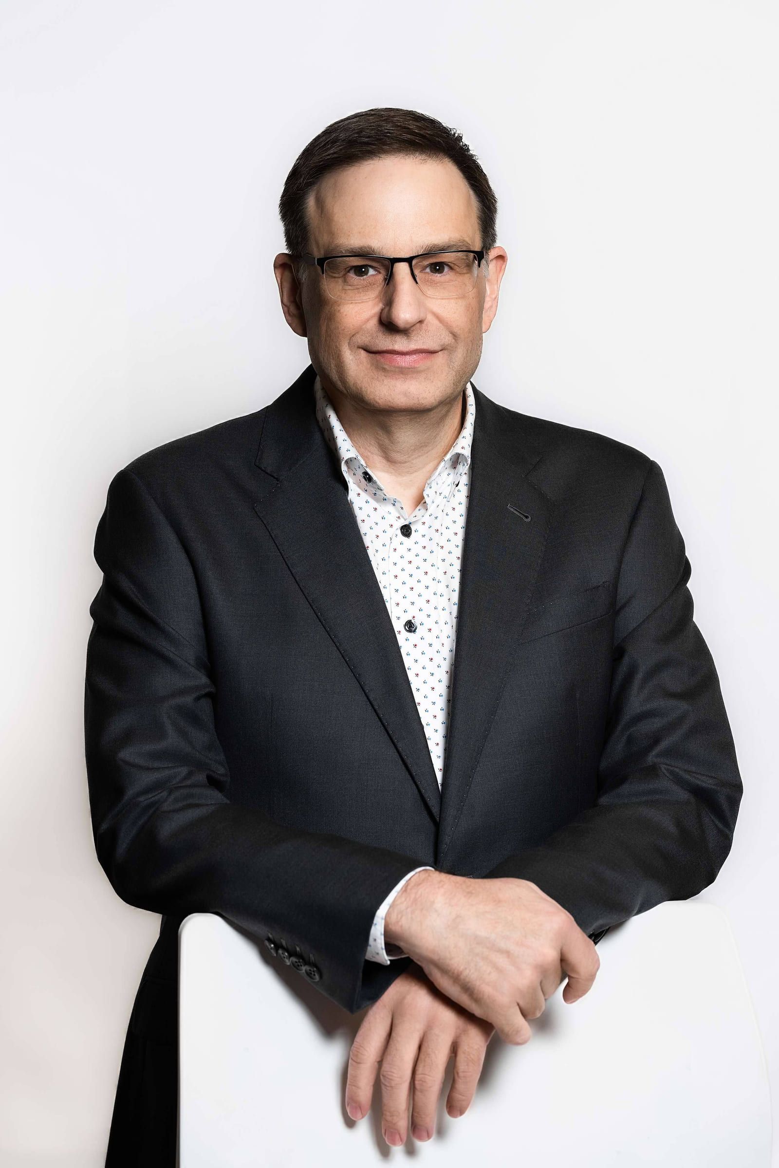 Man in a black suit and glasses leaning on a chair, white backdrop