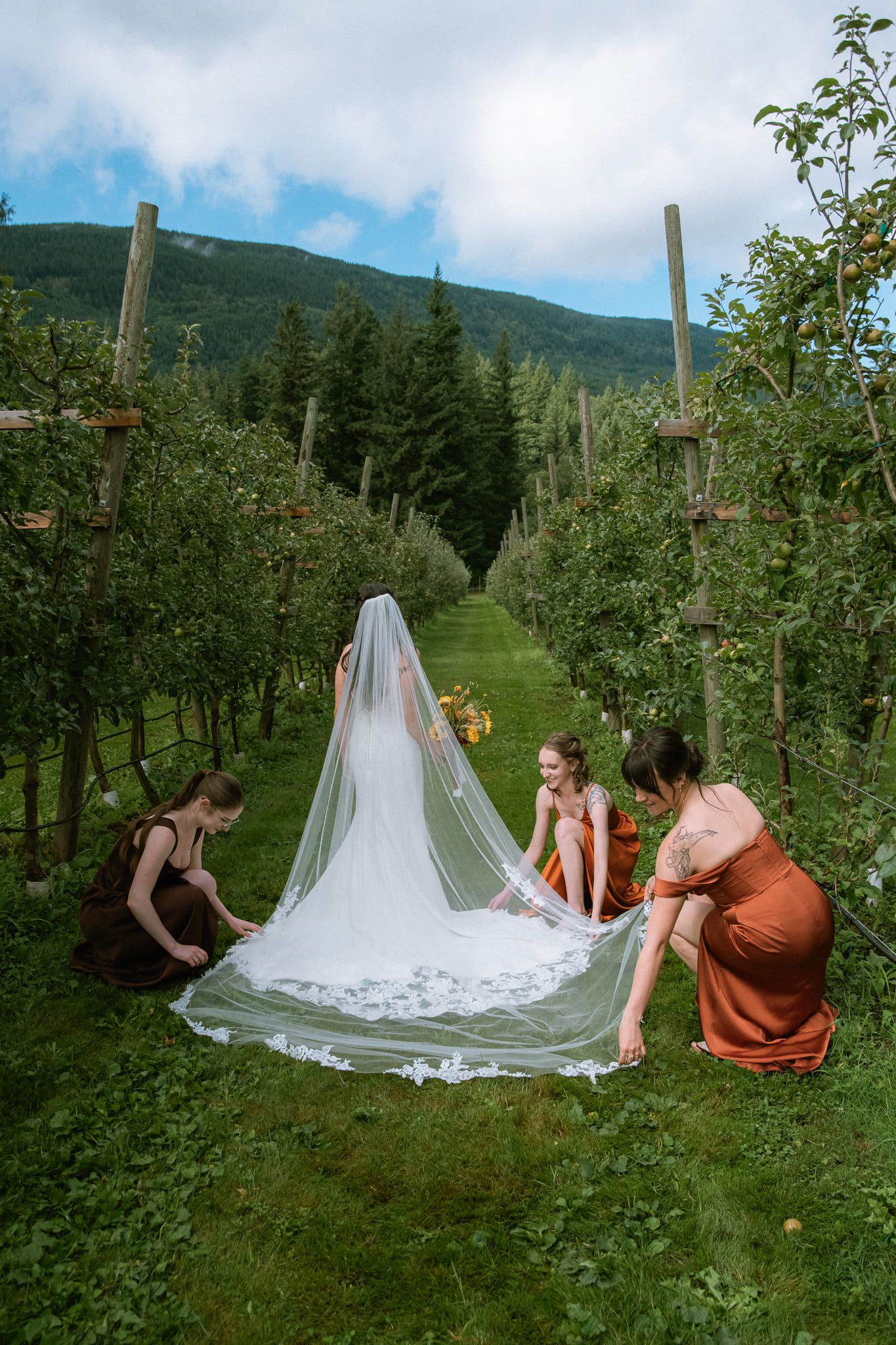 Moody wedding photography, bridesmaids spreading the bride's long veil between rows of an orchard