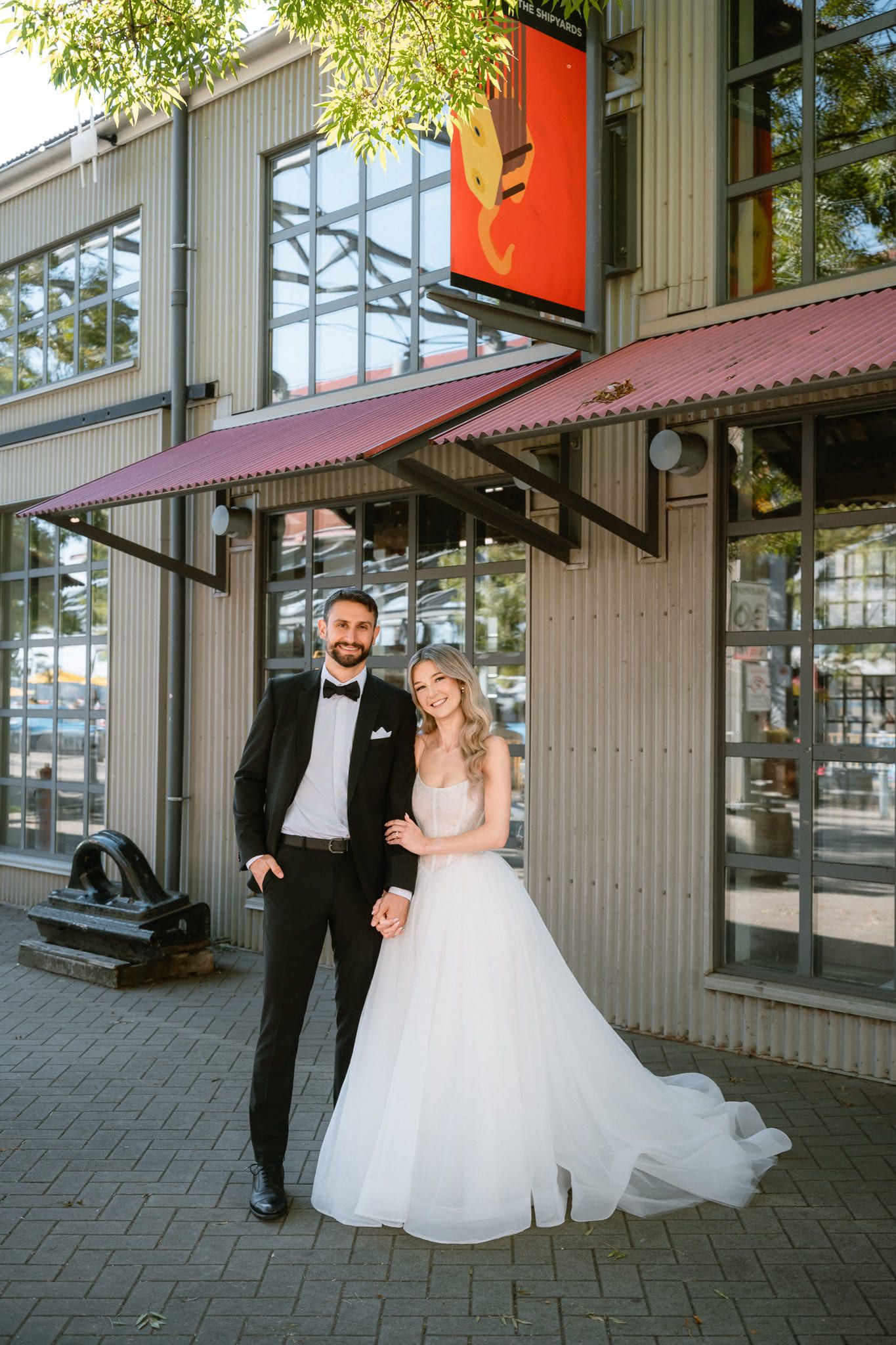 Bride in a tulle gown and groom in a tuxedo hold hands outside a steel building with a red awning, by Vancouver wedding photographer Natalie Varro