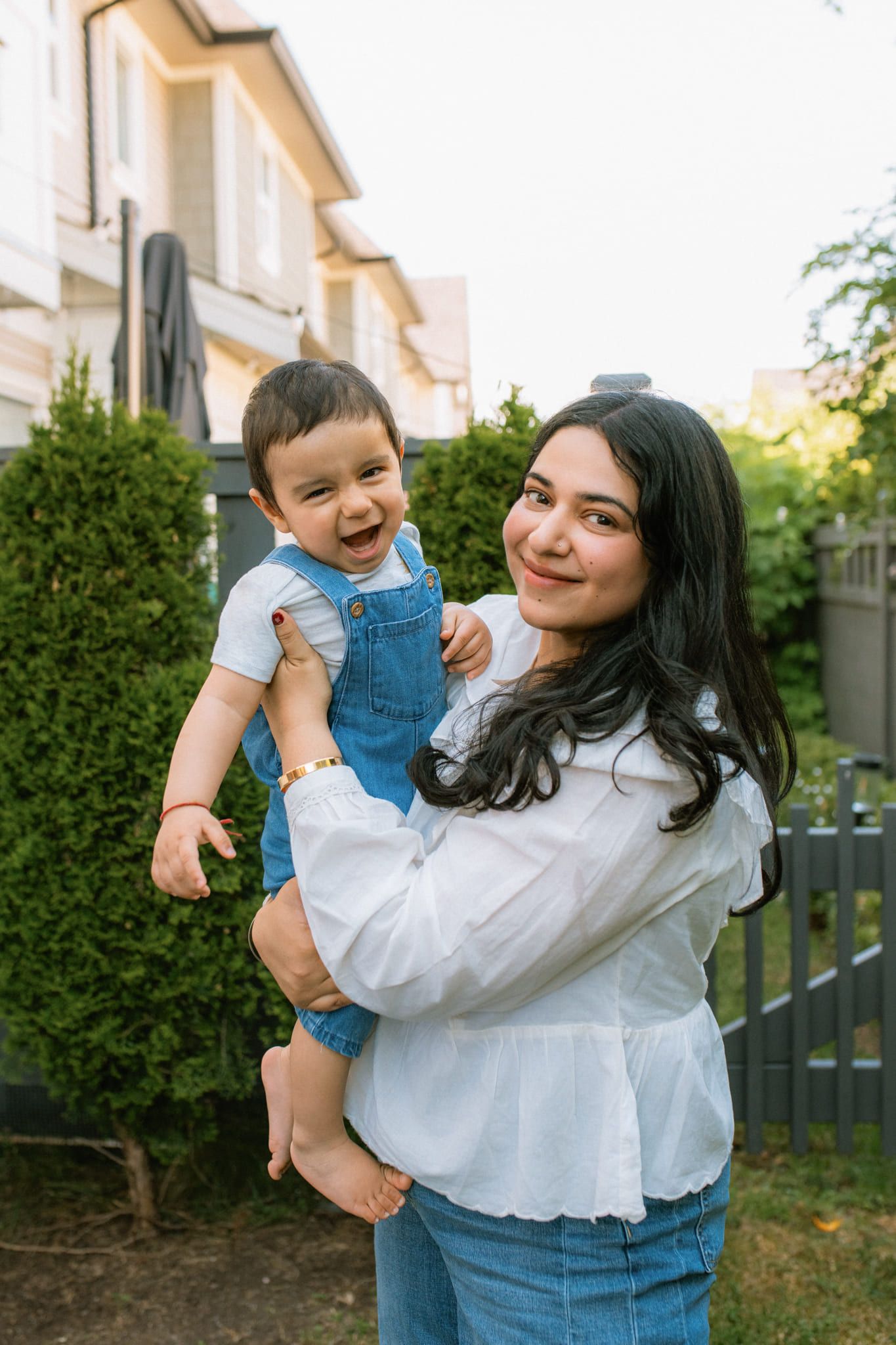Natural family photoshoot, mother holding her laughing toddler in denim overalls outside their home