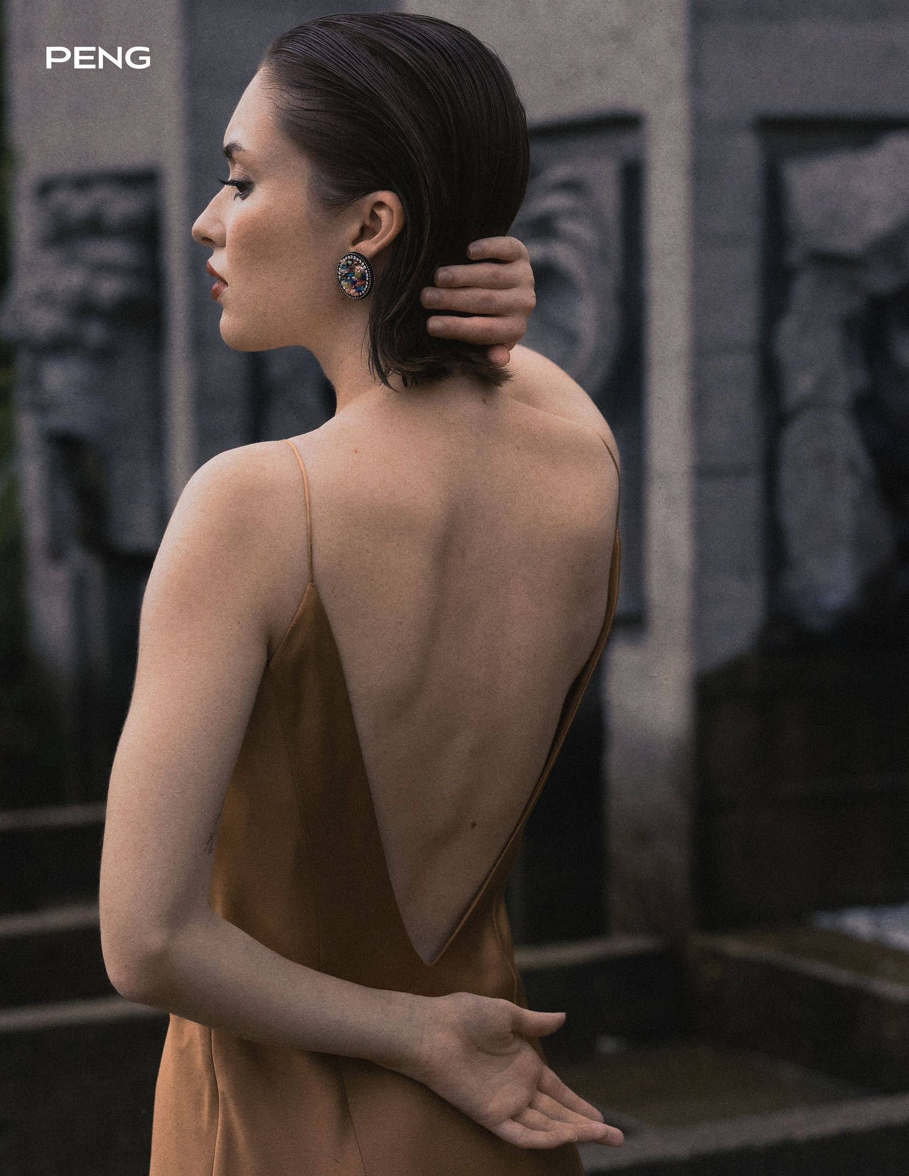 Model in a backless satin dress with a hand in her hair by a stone wall, from PENG magazine