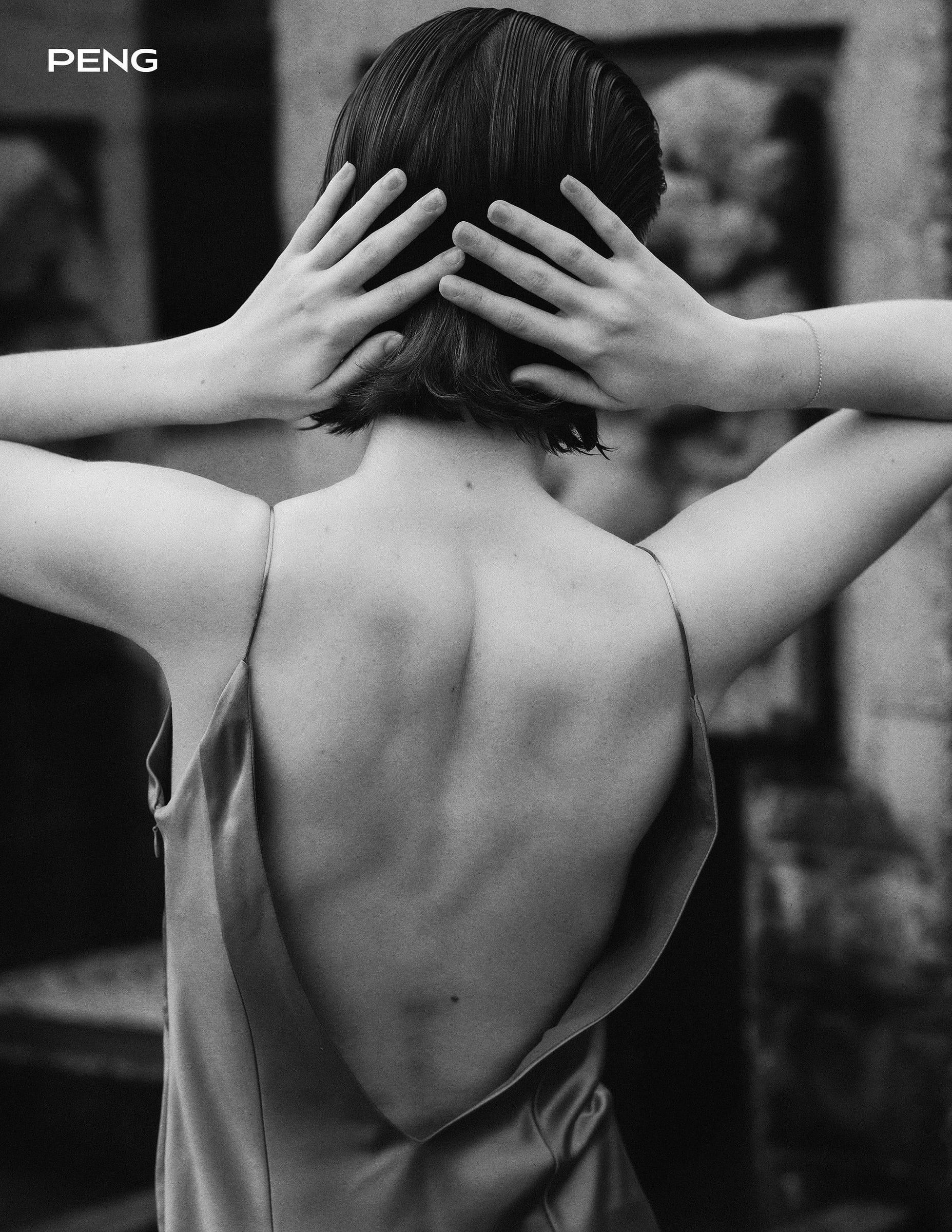 Black-and-white photo of a model in a backless dress covering her face with both hands