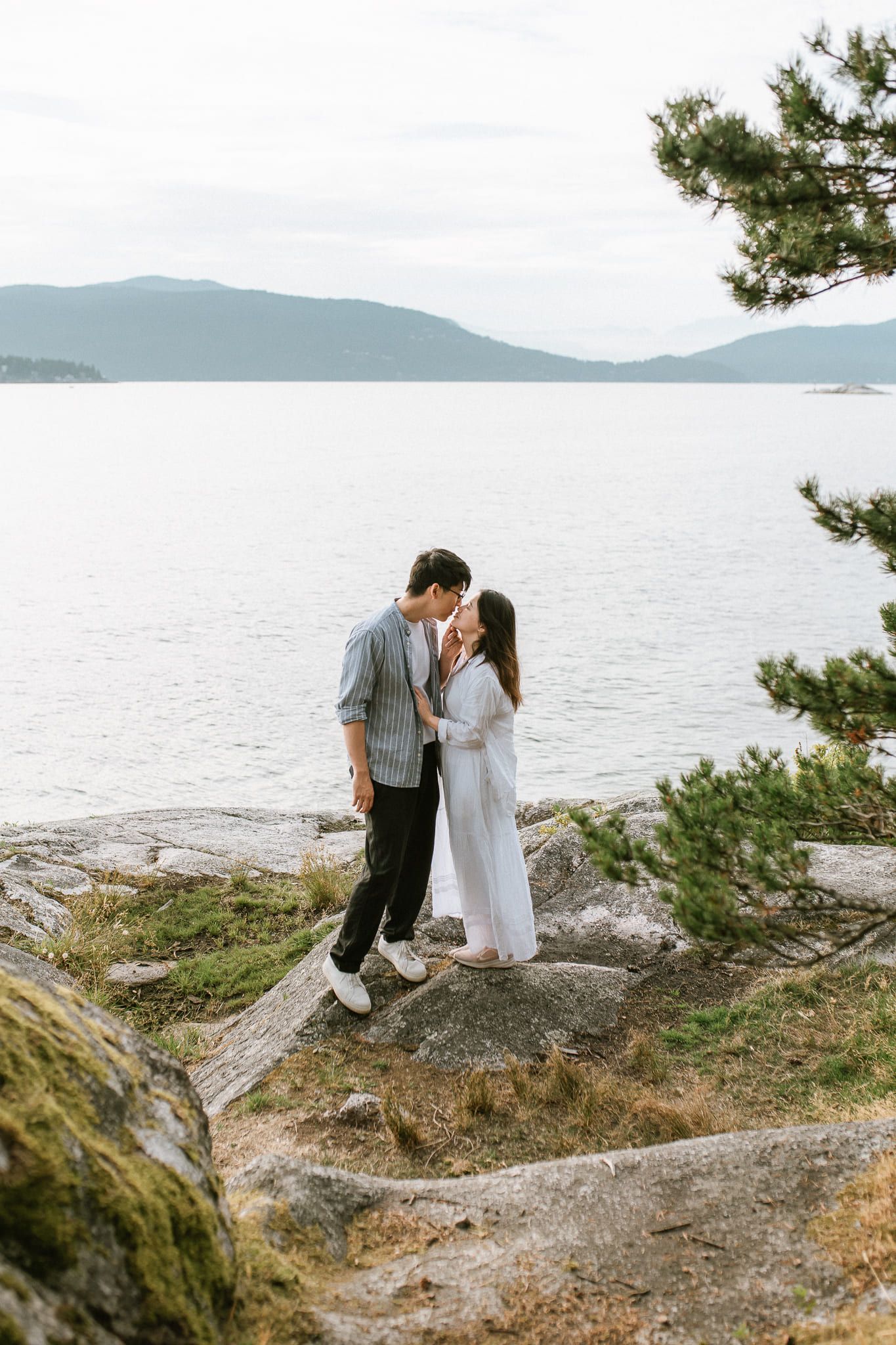 Simple engagement pictures, a couple kiss on a rocky point above the ocean, mountains across the water and a pine tree beside them