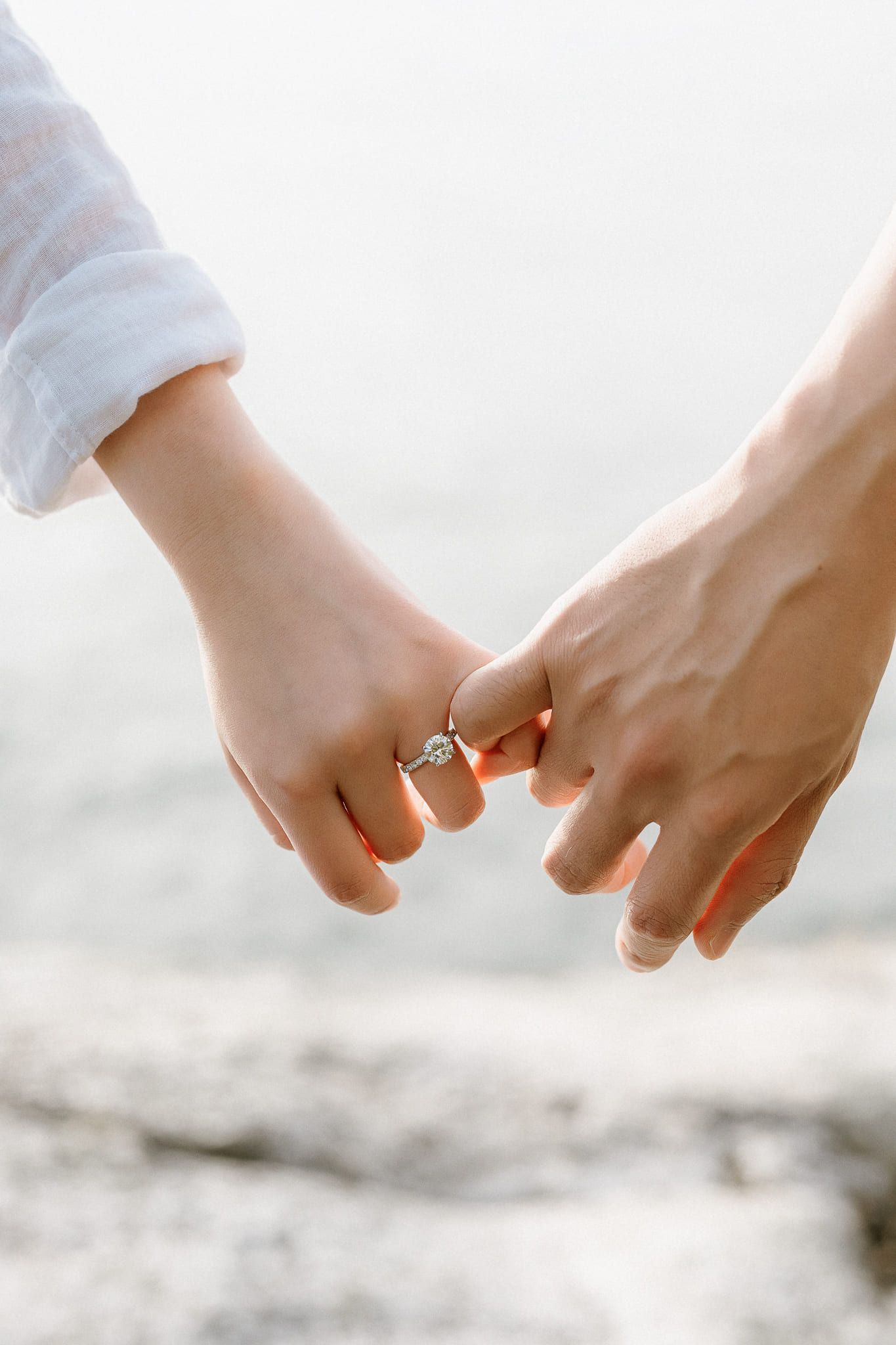 Classic engagement pictures, close-up of two hands linked, a diamond engagement ring on her finger, the sea blurred behind